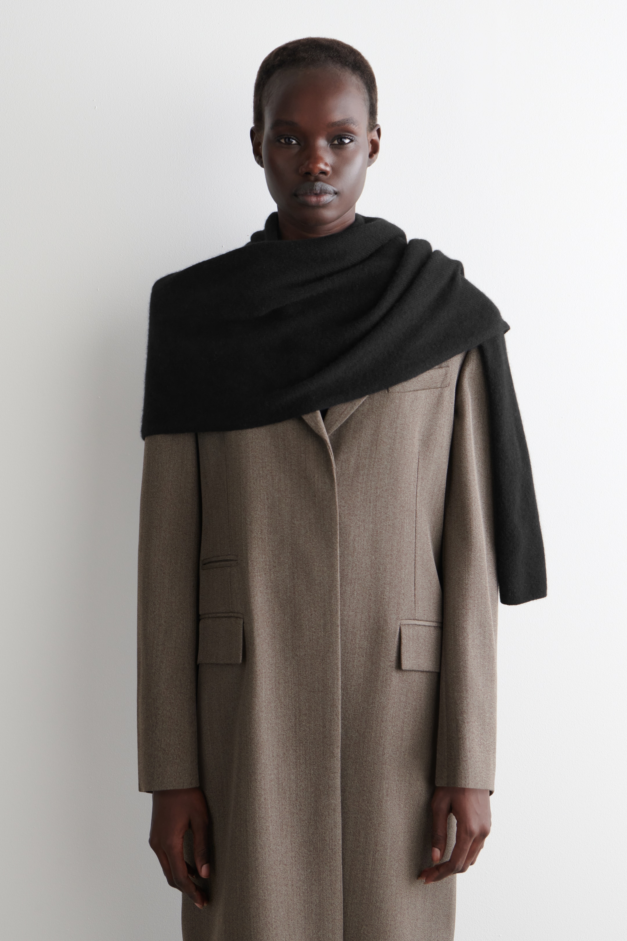 View larger image: A long, muted brown overcoat with a subtle textured weave is being worn, featuring two large flap pockets and partially obscured notch lapels. A black scarf is being draped over the overcoat's shoulders and neck.