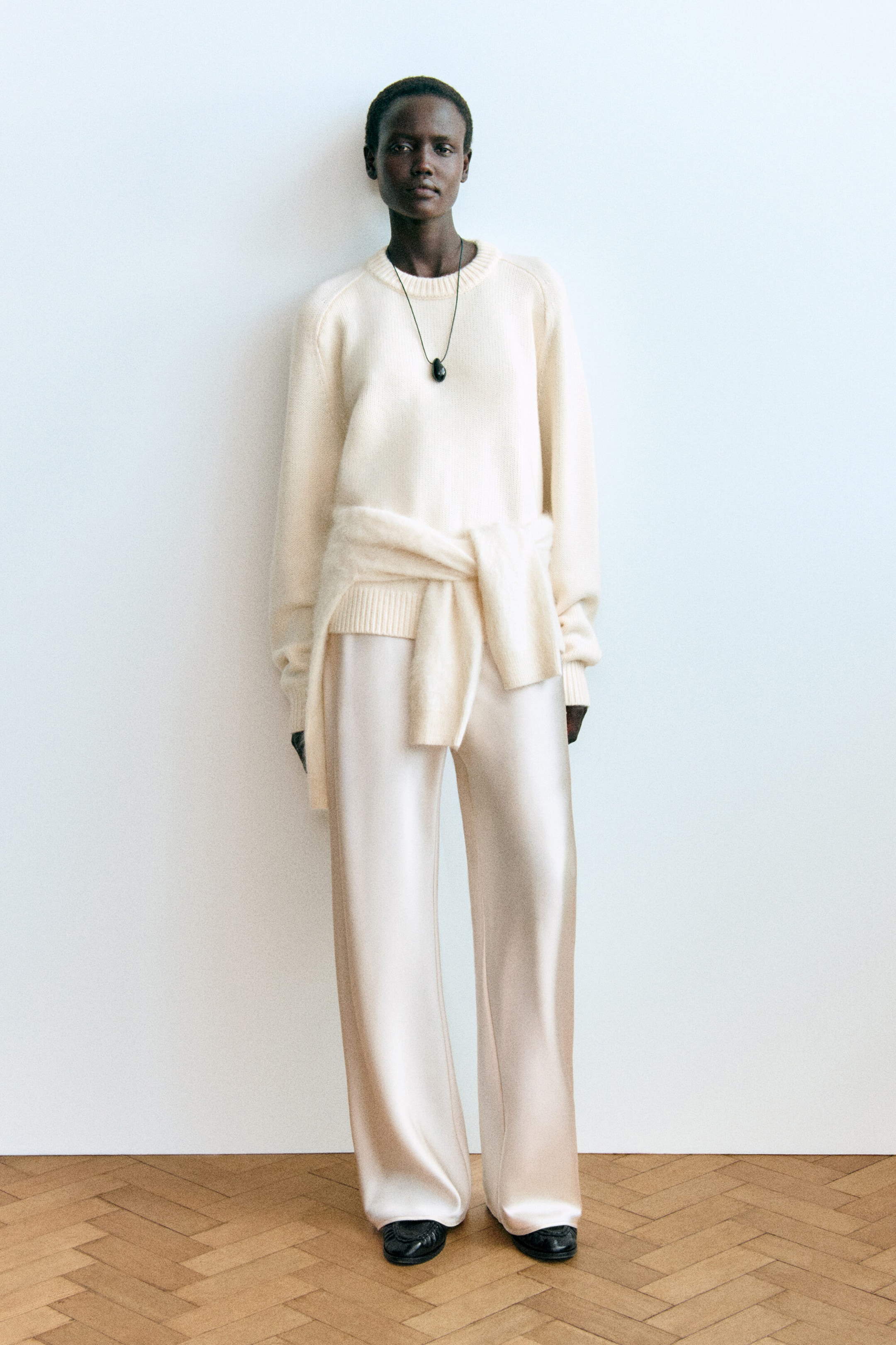 A model is posing front-on, wearing a cream-coloured rounded pure cashmere crew-neck jumper with ribbed trims and champagne-coloured satin wide-leg trousers that feature a fluid drape. Another matching cream knitwear piece is draped around the waist, while a dark pendant necklace is layered over the jumper. A model is posing front-on, wearing a cream-coloured rounded pure cashmere crew-neck jumper with ribbed trims and champagne-coloured satin wide-leg trousers that feature a fluid drape. Another matching cream knitwear piece is draped around the waist, while a dark pendant necklace is layered over the jumper.
