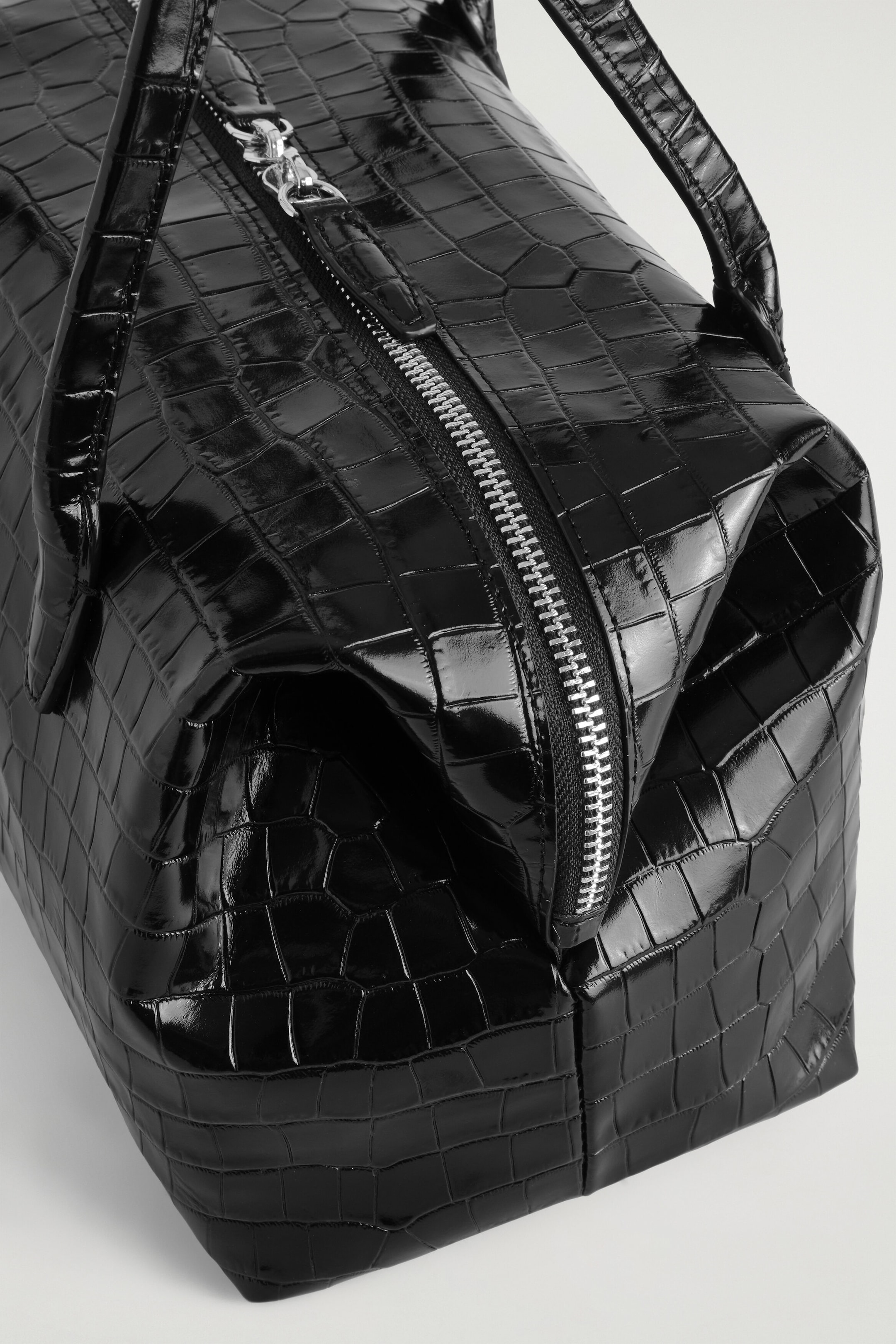 A black glossy leather bowling bag showcases a debossed croc motif and a sculptural shape with softly pinched sides. The bag features a silver-tone two-way zip closure running along the top and down the side, along with matching top handles. A black glossy leather bowling bag showcases a debossed croc motif and a sculptural shape with softly pinched sides. The bag features a silver-tone two-way zip closure running along the top and down the side, along with matching top handles.