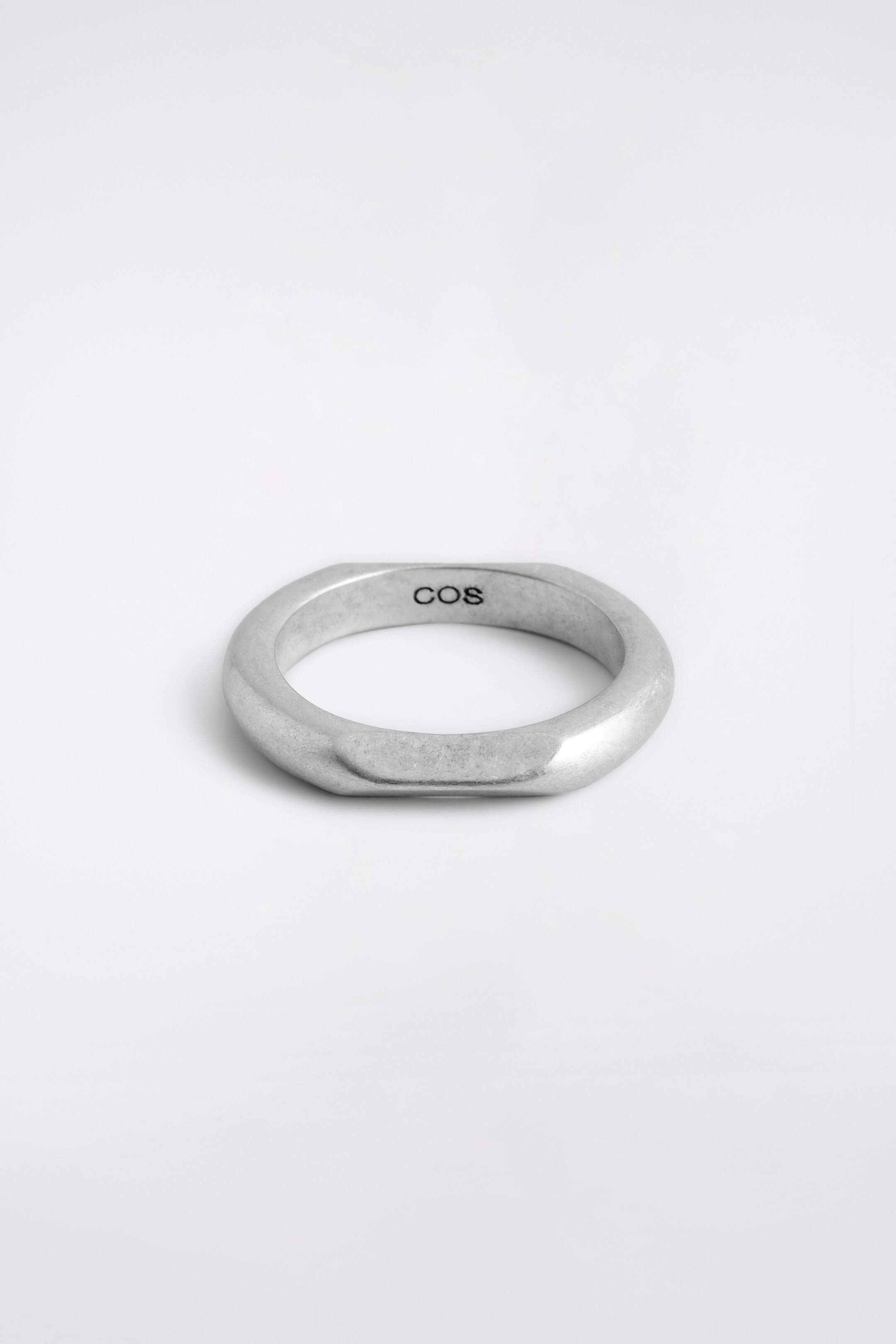 Agrandir l'image: Une bague de ton argenté est exposée à plat, mettant en valeur son anneau épais et irrégulier avec un dessus et un dessous aplatis et des côtés subtilement ondulés et inclinés. Le nom de la marque "COS" est visible sur sa surface intérieure.