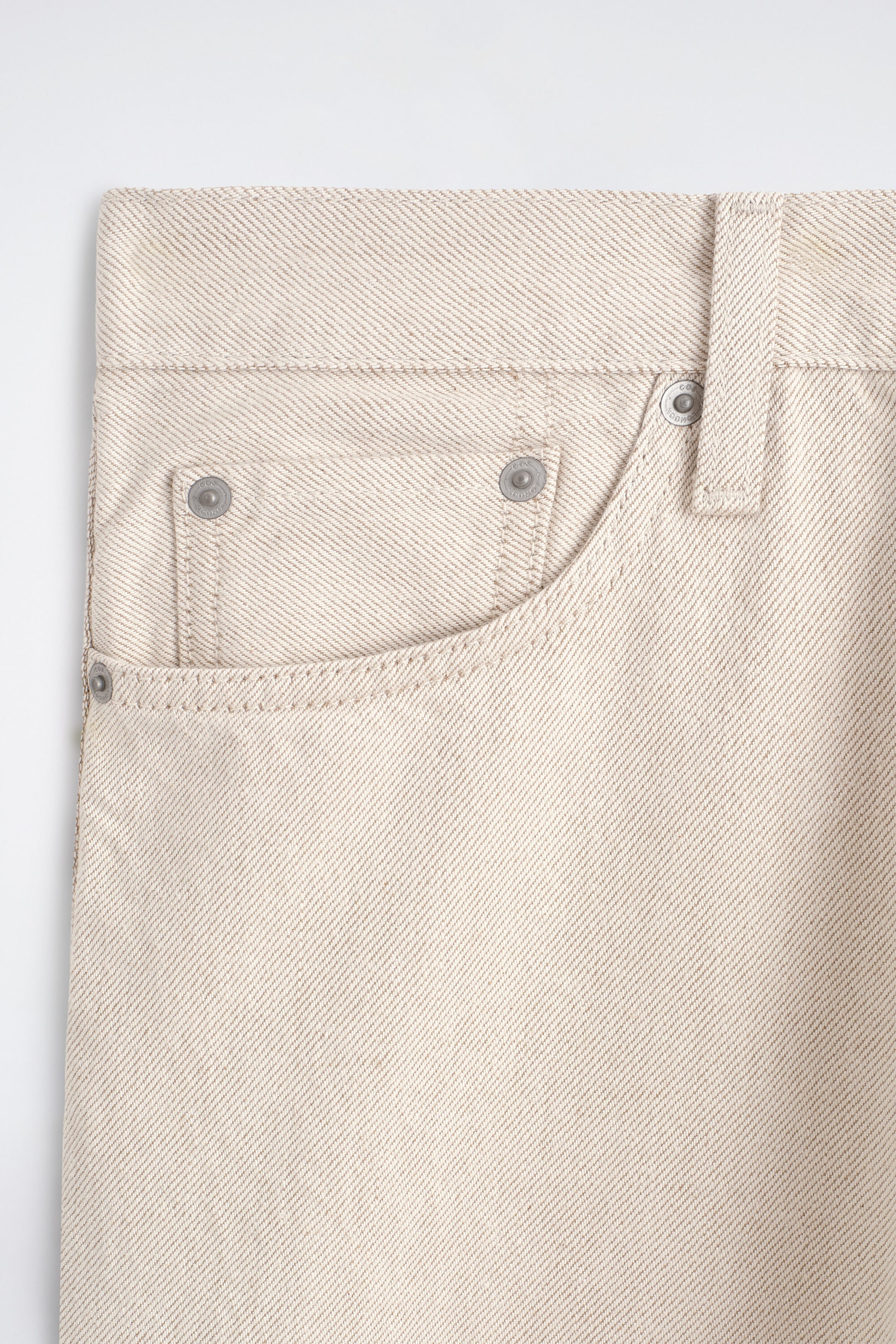 Eine Nahaufnahme der Vorderseite einer hellbeigen Denimhose zeigt den Bund, eine Gürtelschlaufe, eine geschwungene Fronttasche und eine kleine Münztasche, die mit Metallnieten befestigt ist. Die markante diagonale Twill-Struktur des Stoffes und die farblich passende Absteppung sind klar sichtbar. Eine Nahaufnahme der Vorderseite einer hellbeigen Denimhose zeigt den Bund, eine Gürtelschlaufe, eine geschwungene Fronttasche und eine kleine Münztasche, die mit Metallnieten befestigt ist. Die markante diagonale Twill-Struktur des Stoffes und die farblich passende Absteppung sind klar sichtbar.