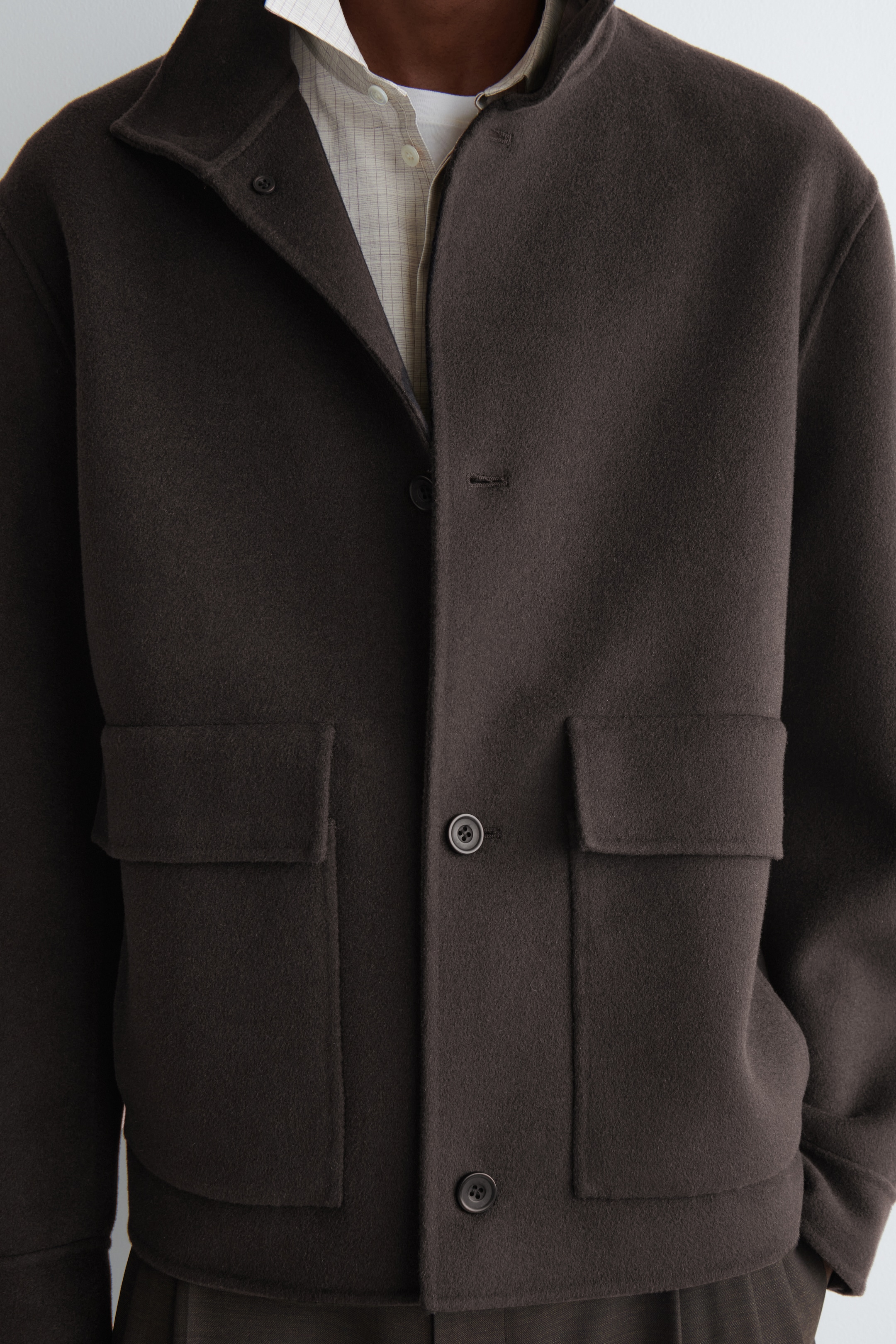 Grösseres Bild ansehen: Eine dunkelbraune Jacke mit einer strukturierten Oberfläche wird von dem Model getragen. Sie verfügt über einen Knopfverschluss, einen spitzen Kragen und zwei große Klappentaschen auf der Vorderseite. Die Jacke ist über einem hellen karierten Hemd geschichtet.