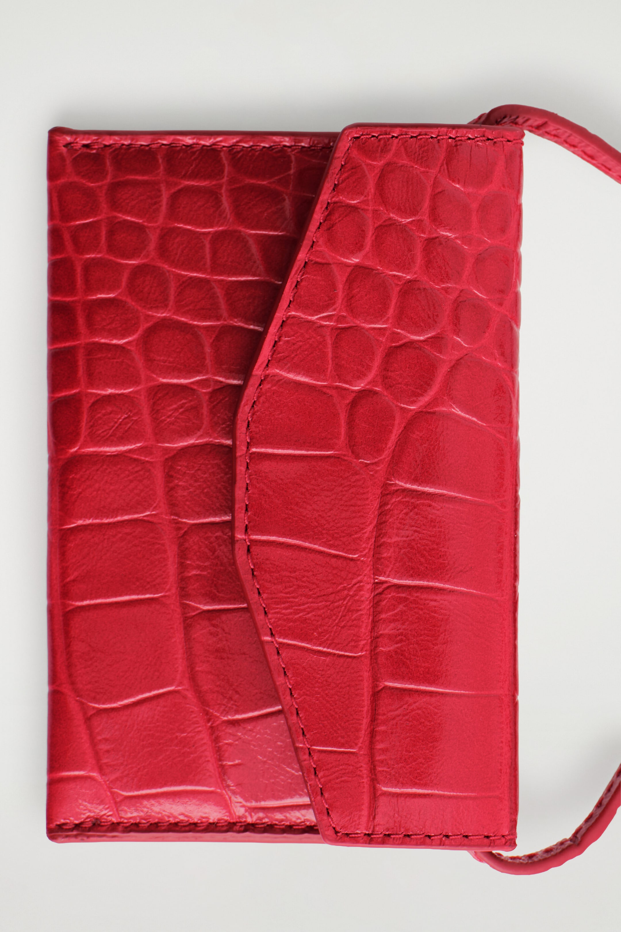 A red leather card holder with a crocodile-embossed texture is shown from the front, showcasing a curved, envelope-style front flap with tonal stitching. A slim, matching red leather strap extends from the side of the compact, rectangular design. A red leather card holder with a crocodile-embossed texture is shown from the front, showcasing a curved, envelope-style front flap with tonal stitching. A slim, matching red leather strap extends from the side of the compact, rectangular design.