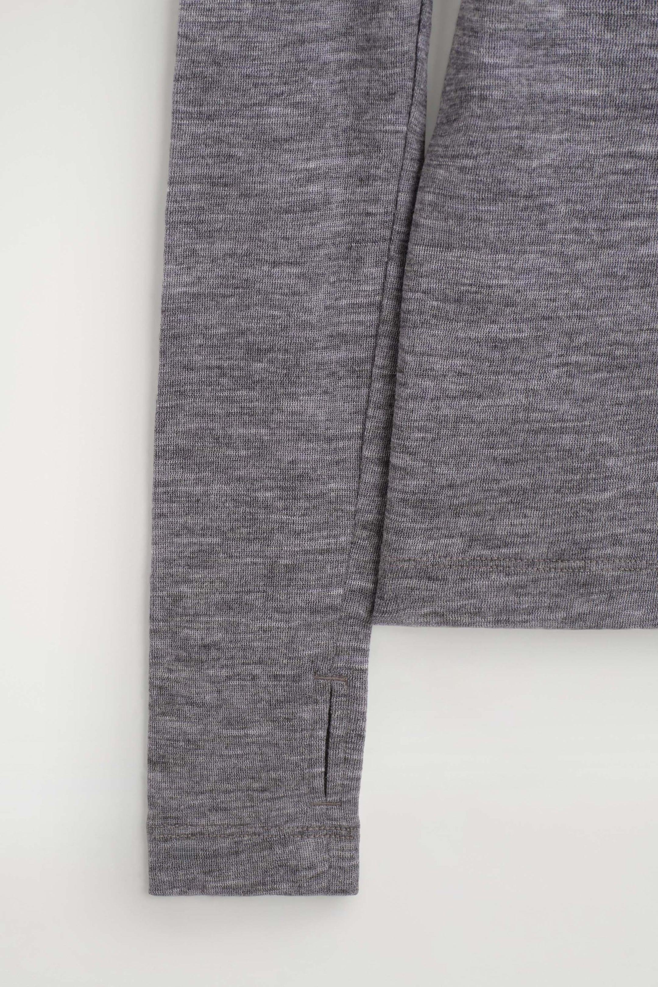 The slim merino wool top features a grey heathered knit fabric, showing a close-up of a long sleeve with a thumbhole cuff alongside the straight hem of the body.