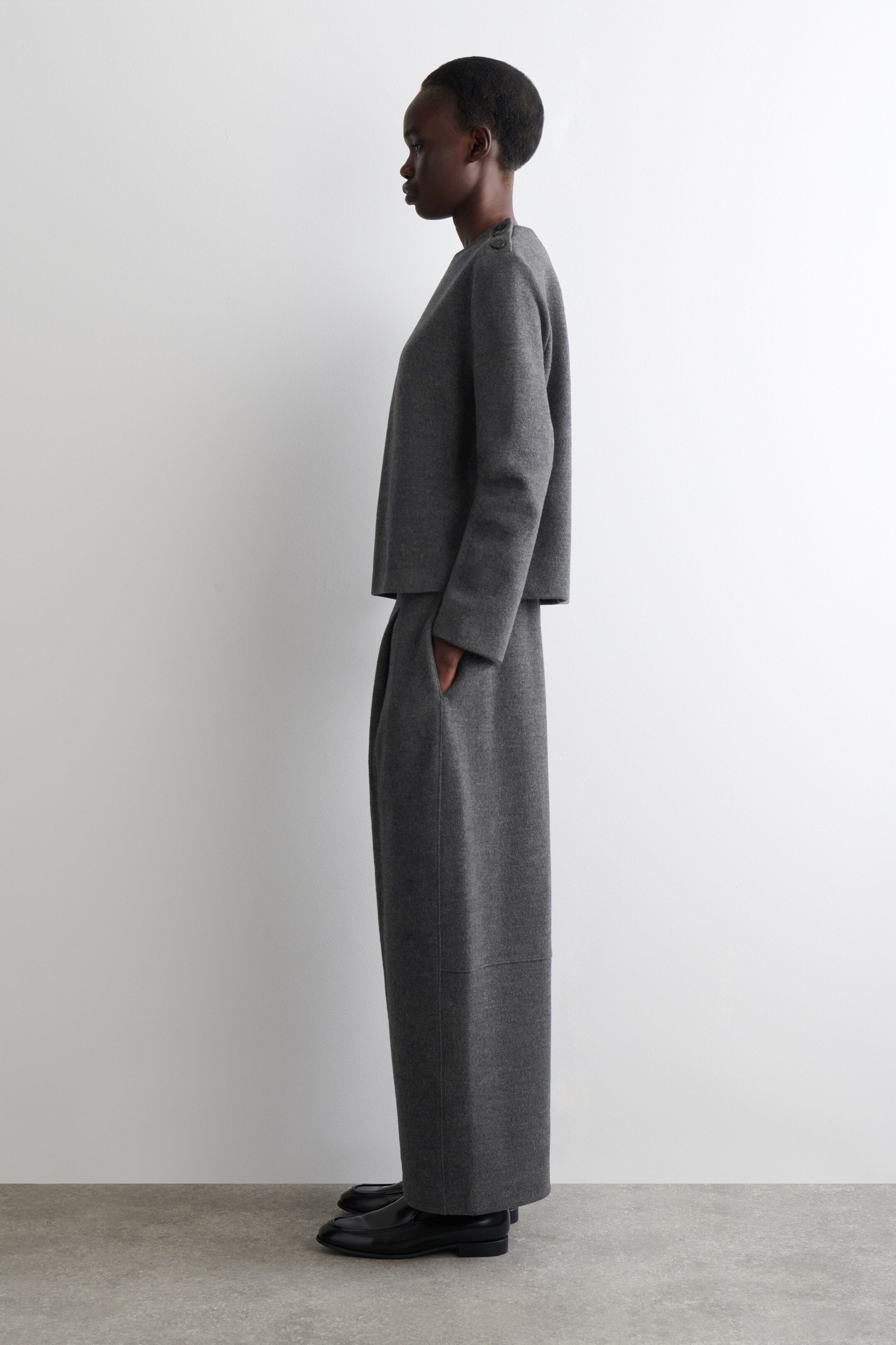 Agrandir l'image: Une tenue deux pièces gris foncé est présentée en profil gauche, avec un haut court et carré doté d'un col rond et de deux boutons sur l'épaule gauche. Le pantalon assorti à jambes larges s'étend jusqu'à la pleine longueur, avec une main reposant dans une poche latérale, et est stylé avec des chaussures noires à enfiler.