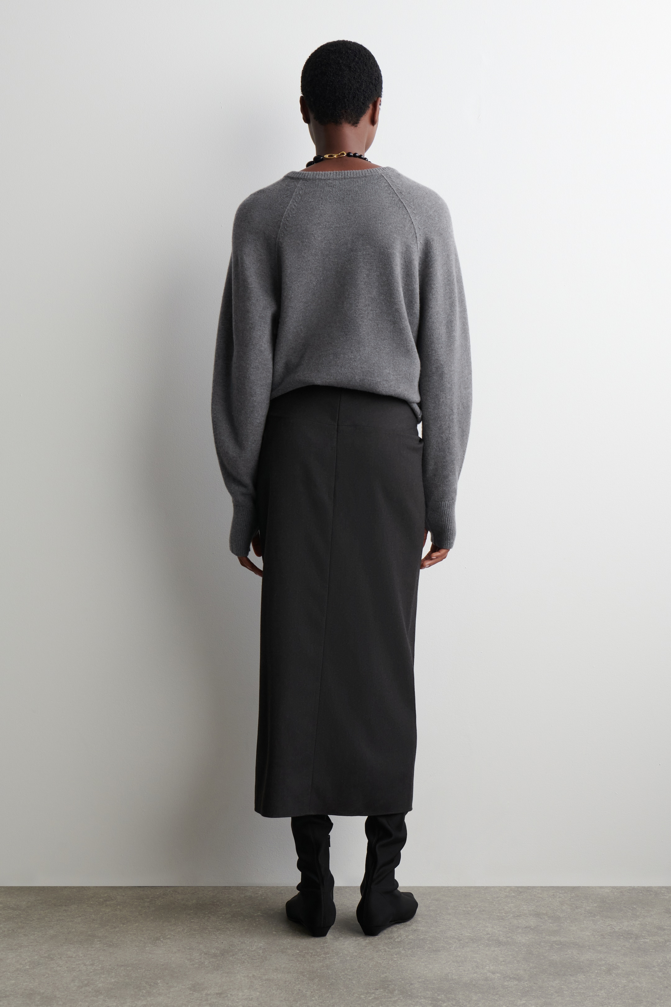 Nagyobb kép megtekintése: A DRAPED WOOL-BLEND MIDI SKIRT van a modellen, aki annak hátulnézetét mutatja be. A fekete midi szoknya térd alá ér, és egy szürke hosszú ujjú pulóverrel és fekete bokacsizmával van kombinálva.