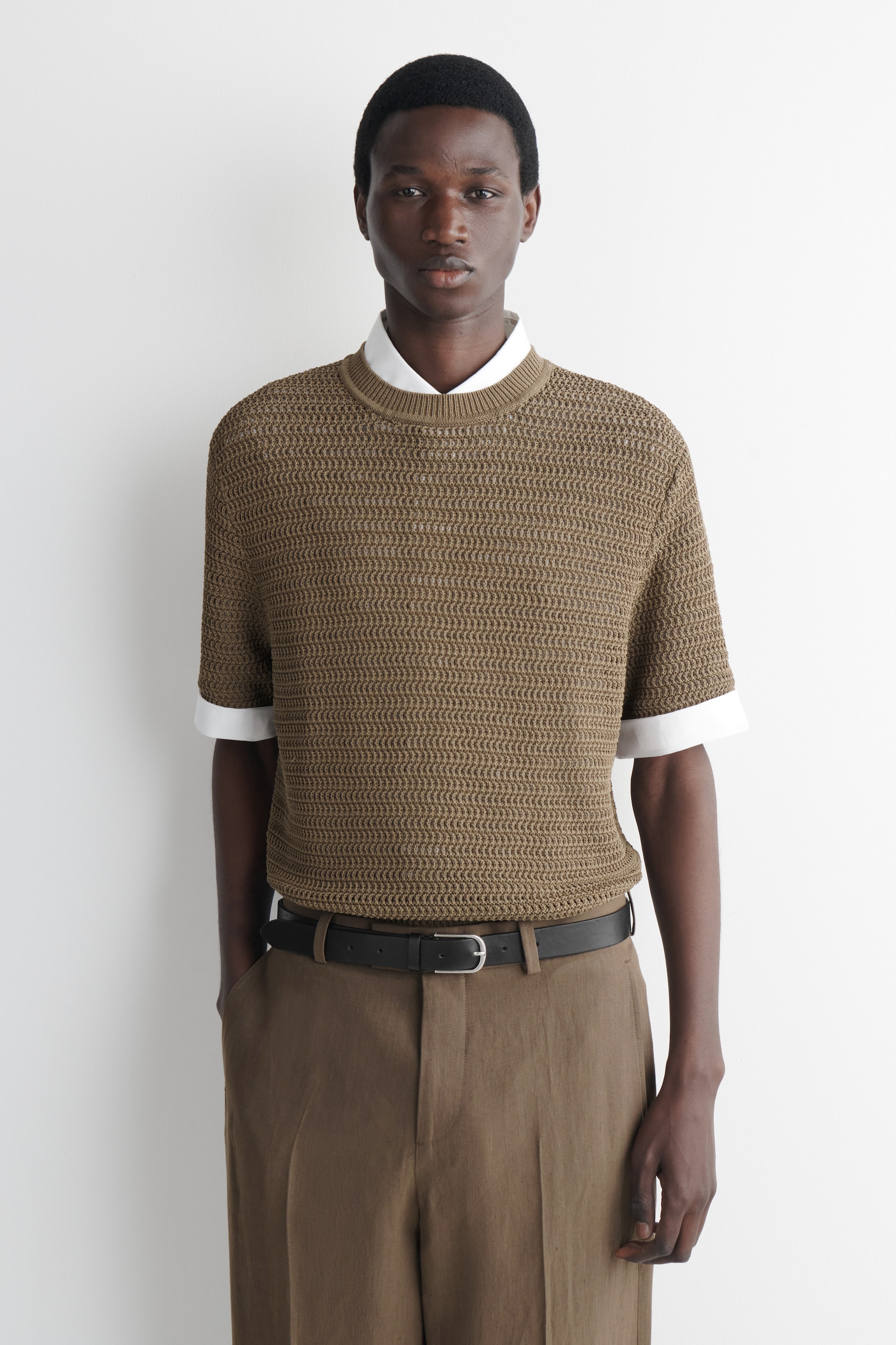 Visualizza immagine più grande: Una T-shirt a maglia aperta in tonalità talpa, con maniche corte e girocollo e bordi a coste, è indossata dalla modella. È stratificata sopra una camicia bianca con colletto e abbinata a pantaloni marroni e una cintura nera.