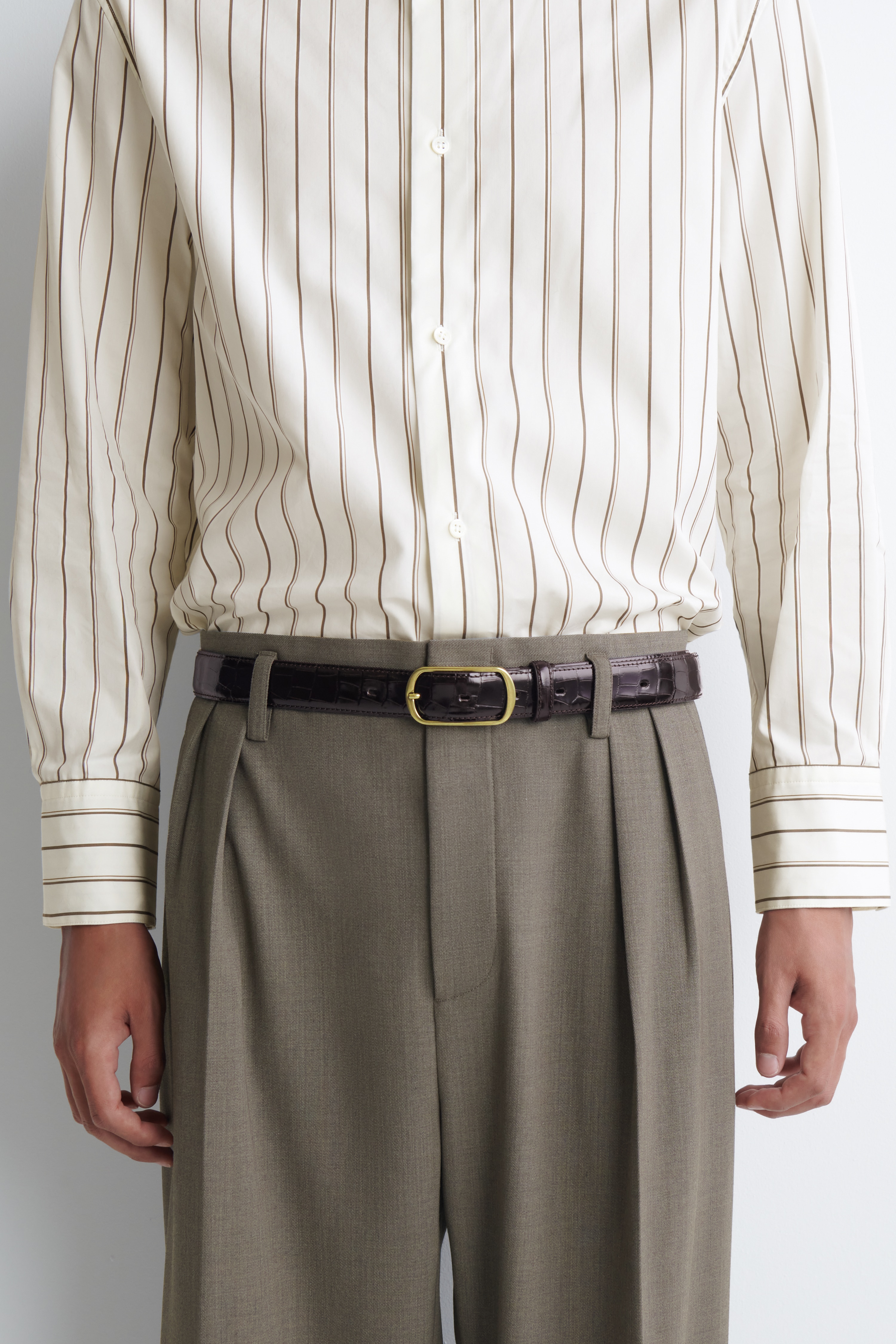 The curved-buckle leather belt is resting in a closed loop, displaying its dark brown, crocodile-embossed leather strap with a glossy finish. It features a gold-toned, rounded rectangular buckle at the front with a single prong and a matching leather keeper.