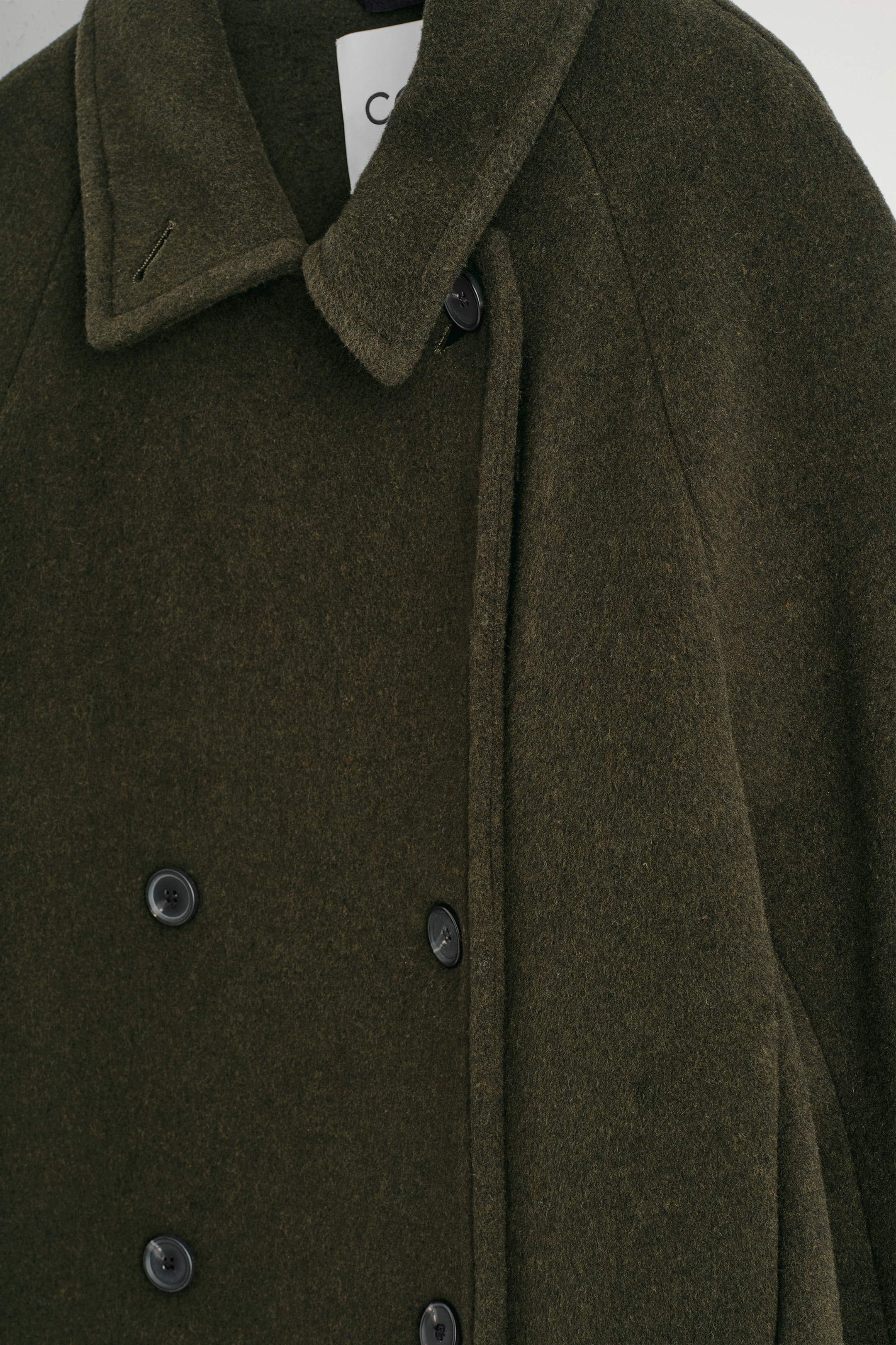 Ingrandisci l'immagine: Un cappotto doppiopetto verde scuro è mostrato dal DAVANTI in un primo piano, evidenziando il suo tessuto strutturato, un colletto in piedi e bottoni scuri sulla chiusura anteriore.