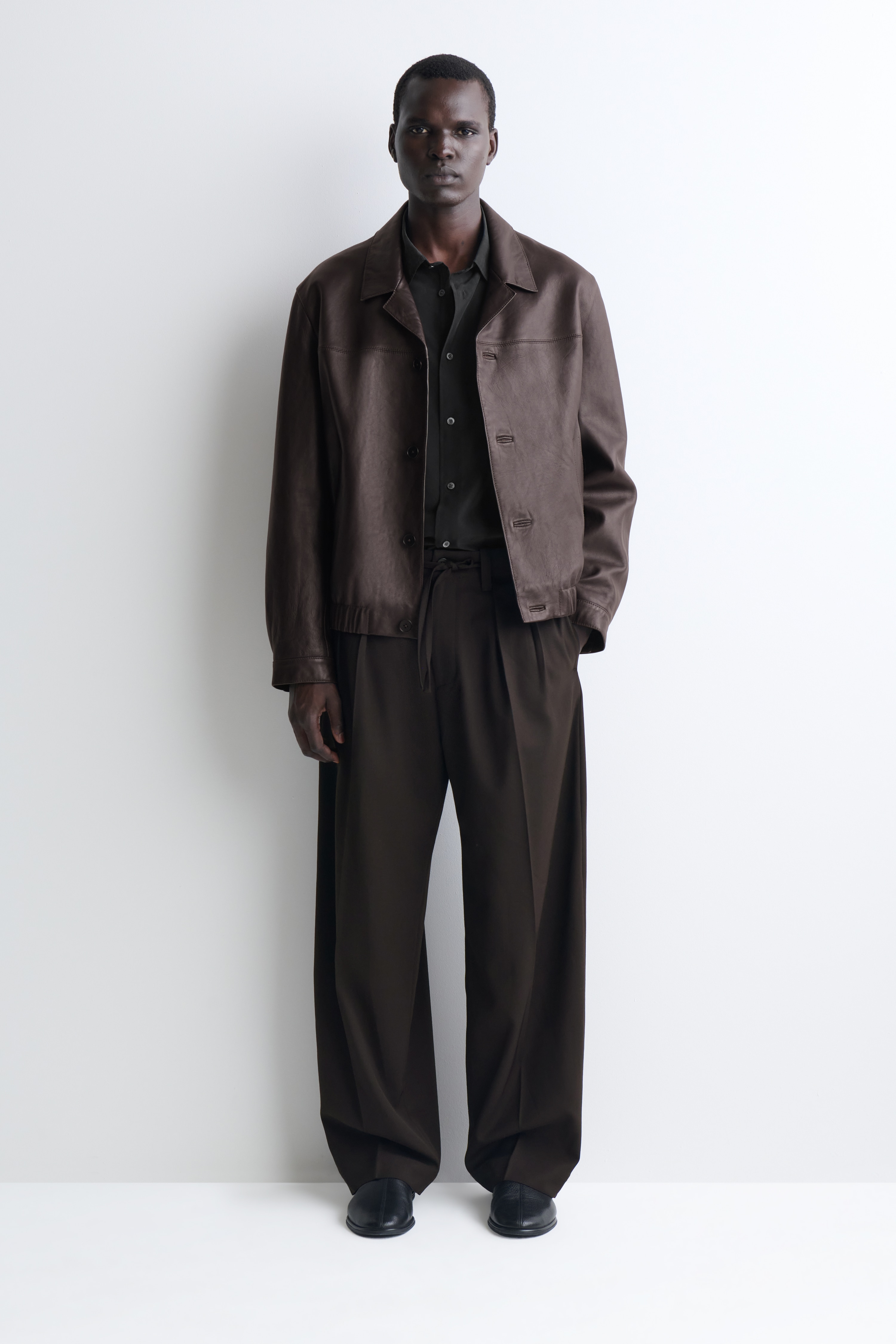 Un paio di pantaloni a gamba larga marrone scuro Ã¨ mostrato frontalmente, con vestibilitÃ  rilassata, singola piega frontale e coulisse in vita coordinata. I pantaloni sono progettati con chiusura con un solo bottone, passanti per cintura e orli dritti.