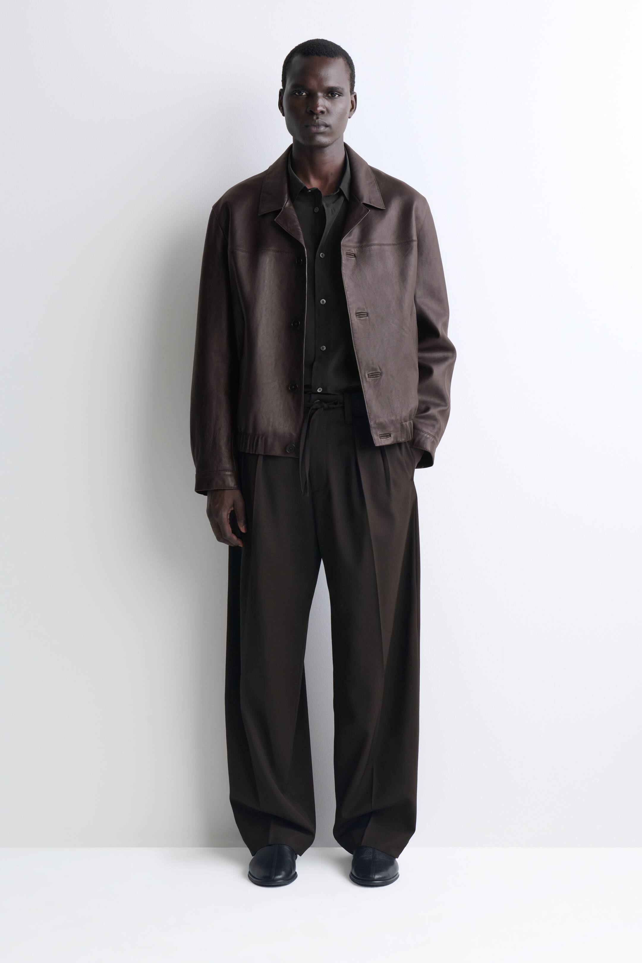 Agrandir l'image: Un modèle se tient de face, portant une veste en cuir marron foncé dotée d’un col à revers, de fermetures boutonnées et d’une taille élastiquée. La veste est portée sur une chemise boutonnée gris charbon et associée à un pantalon marron foncé ample à jambes larges.