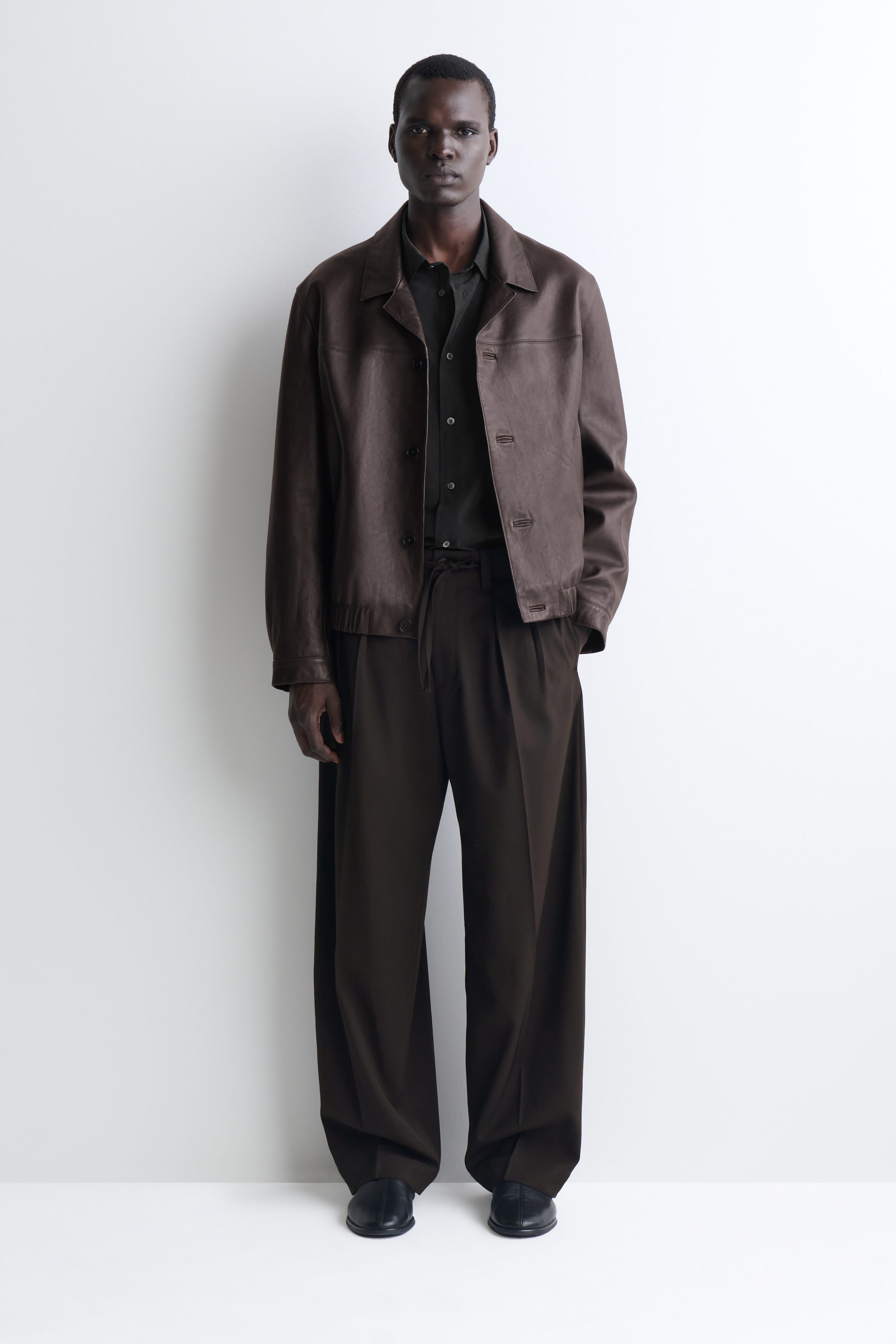 A model is standing front-facing, wearing a dark brown leather jacket with a spread collar, button closures, and an elasticized waistband. The jacket is layered over a charcoal grey button-up shirt and styled with loose, wide-leg dark brown trousers. A model is standing front-facing, wearing a dark brown leather jacket with a spread collar, button closures, and an elasticized waistband. The jacket is layered over a charcoal grey button-up shirt and styled with loose, wide-leg dark brown trousers.