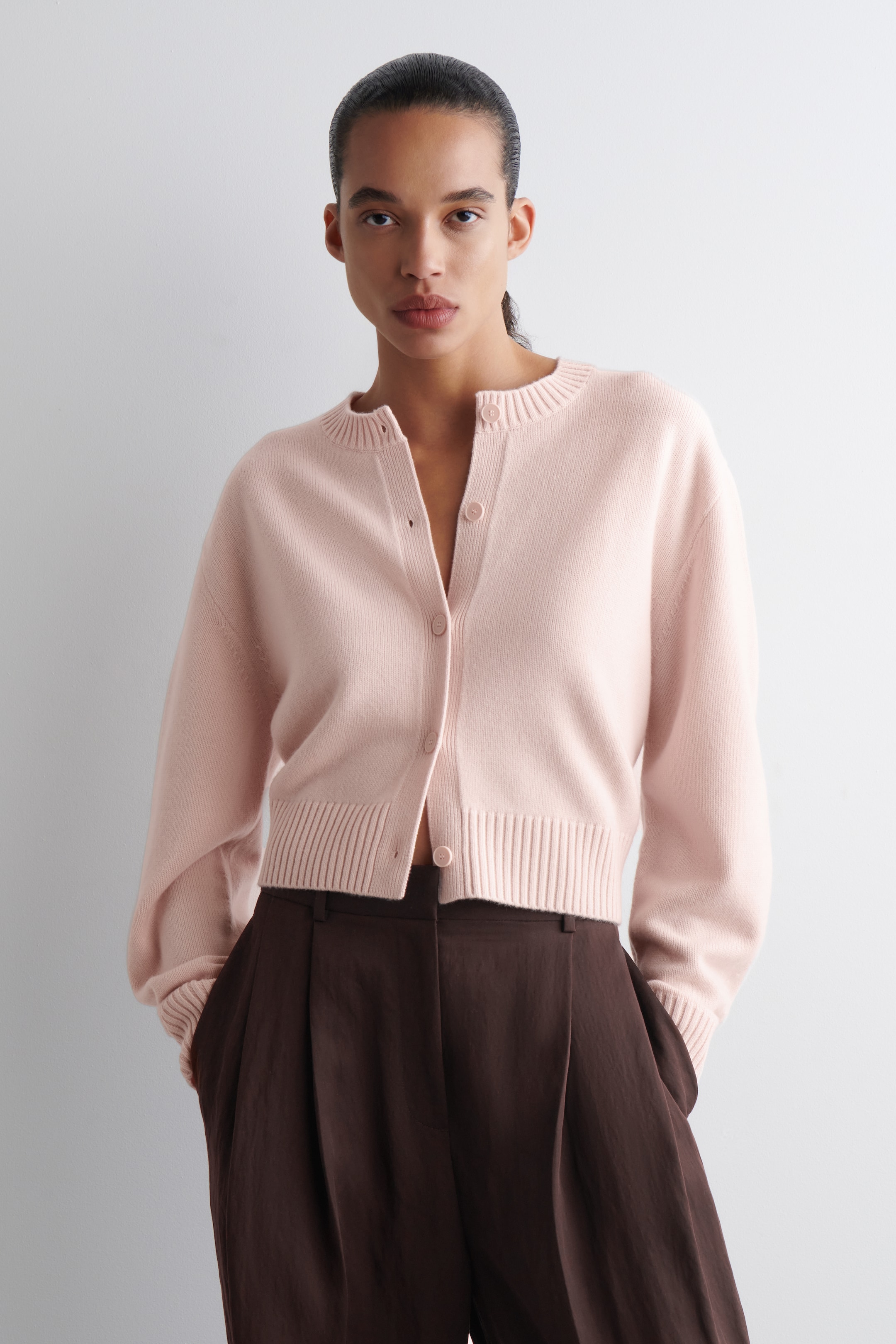 Agrandir l'image: Un cardigan court rose clair est présenté de face, mettant en valeur un col rond, des poignets côtelés et un ourlet côtelé. Il présente une fermeture à boutons sur le devant avec des boutons assortis à la couleur, et sa coupe décontractée ainsi que sa longueur courte sont mises en avant alors qu'il est porté sur un pantalon marron foncé.