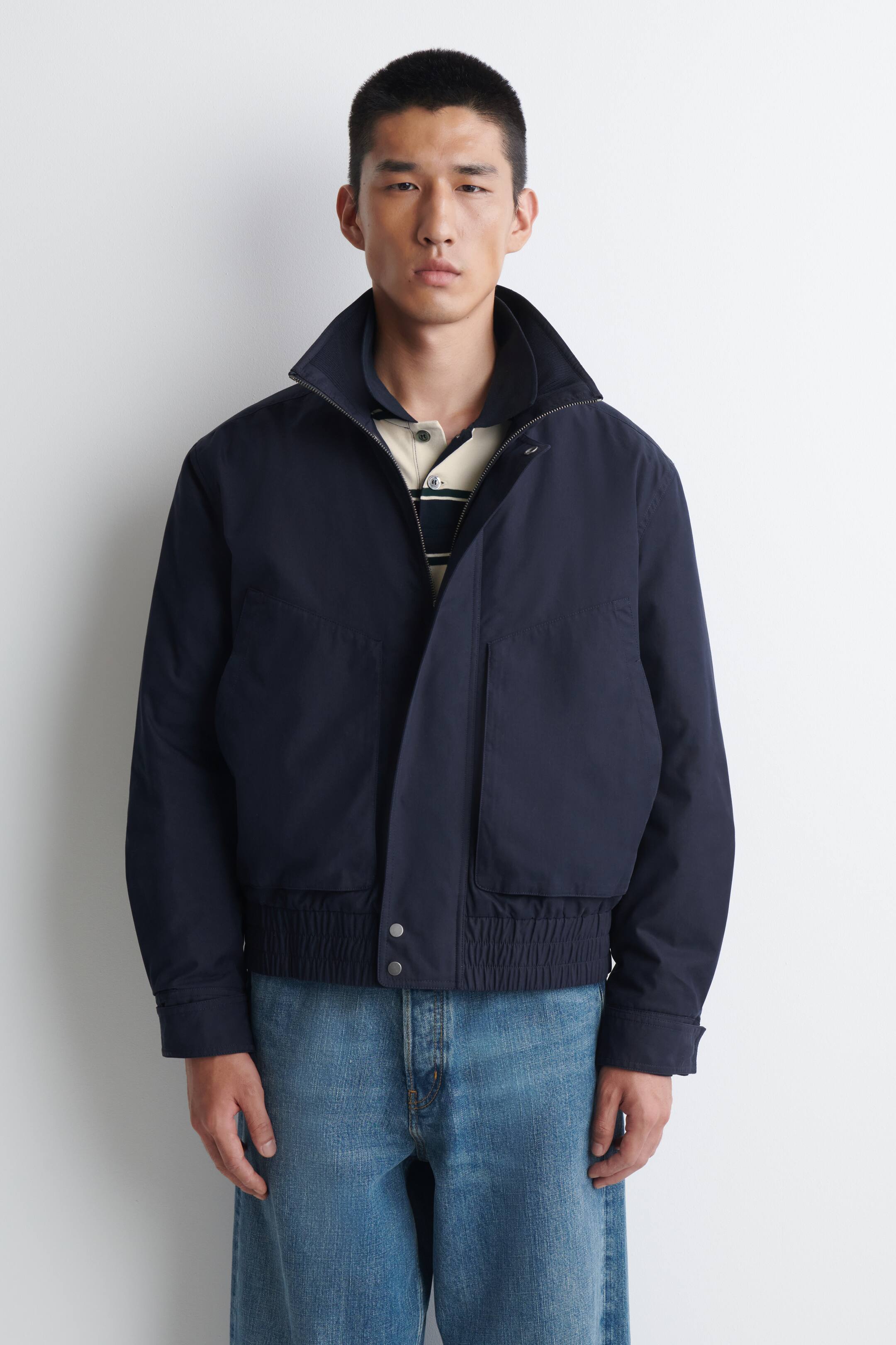 Agrandir l'image: Une veste bomber bleu marine est présentée de face. Elle présente un col montant avec une fermeture éclair visible, deux poches à rabat à l’avant en bas et un ourlet élastiqué. La veste est partiellement ouverte, laissant apparaître un polo rayé en dessous.
