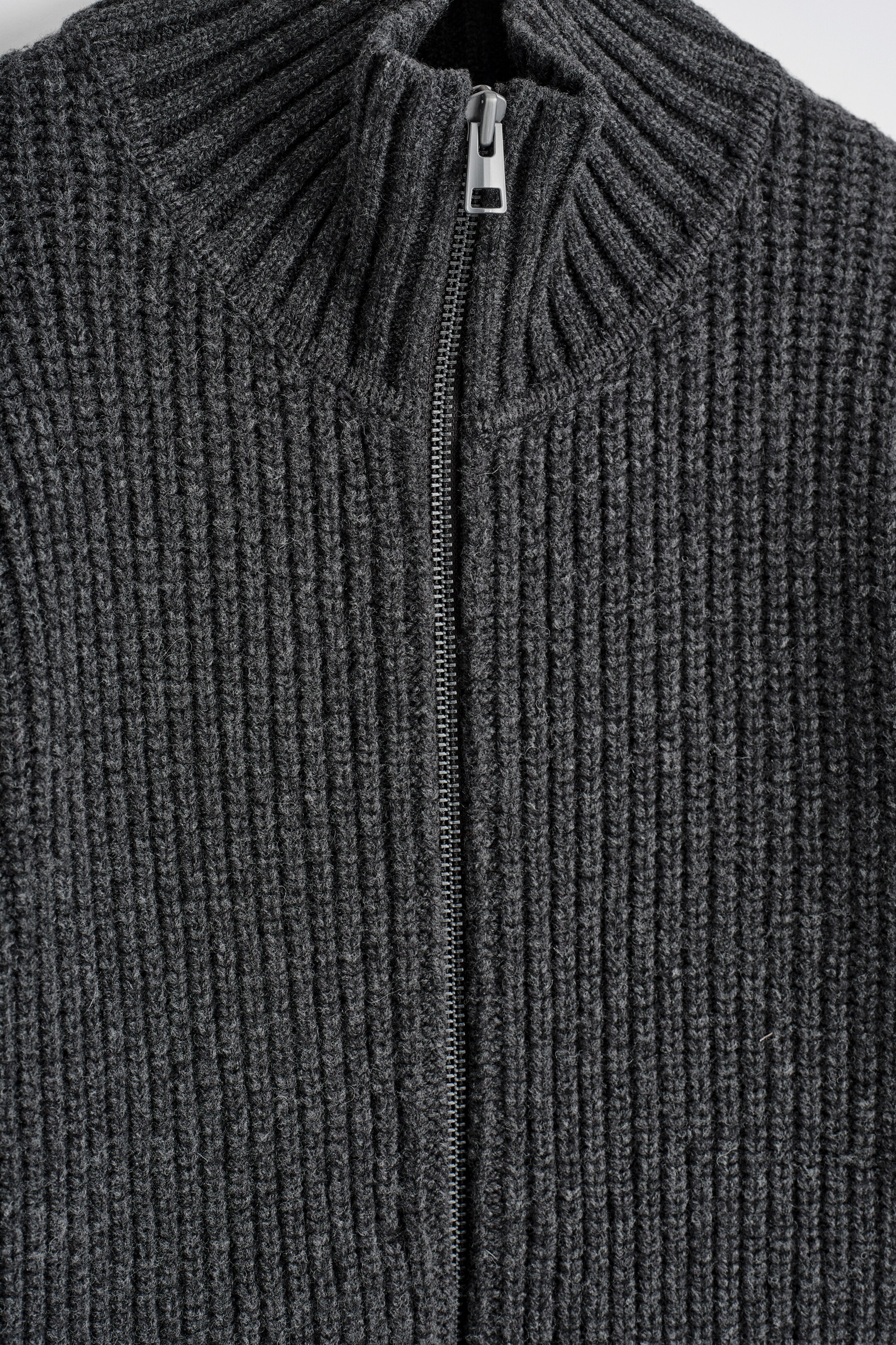 Agrandir l'image: Une veste en tricot gris foncé présente une texture côtelée sur le DEVANT, avec une fermeture éclair de couleur argentée qui s'étend jusqu'à un col cheminée côtelé.
