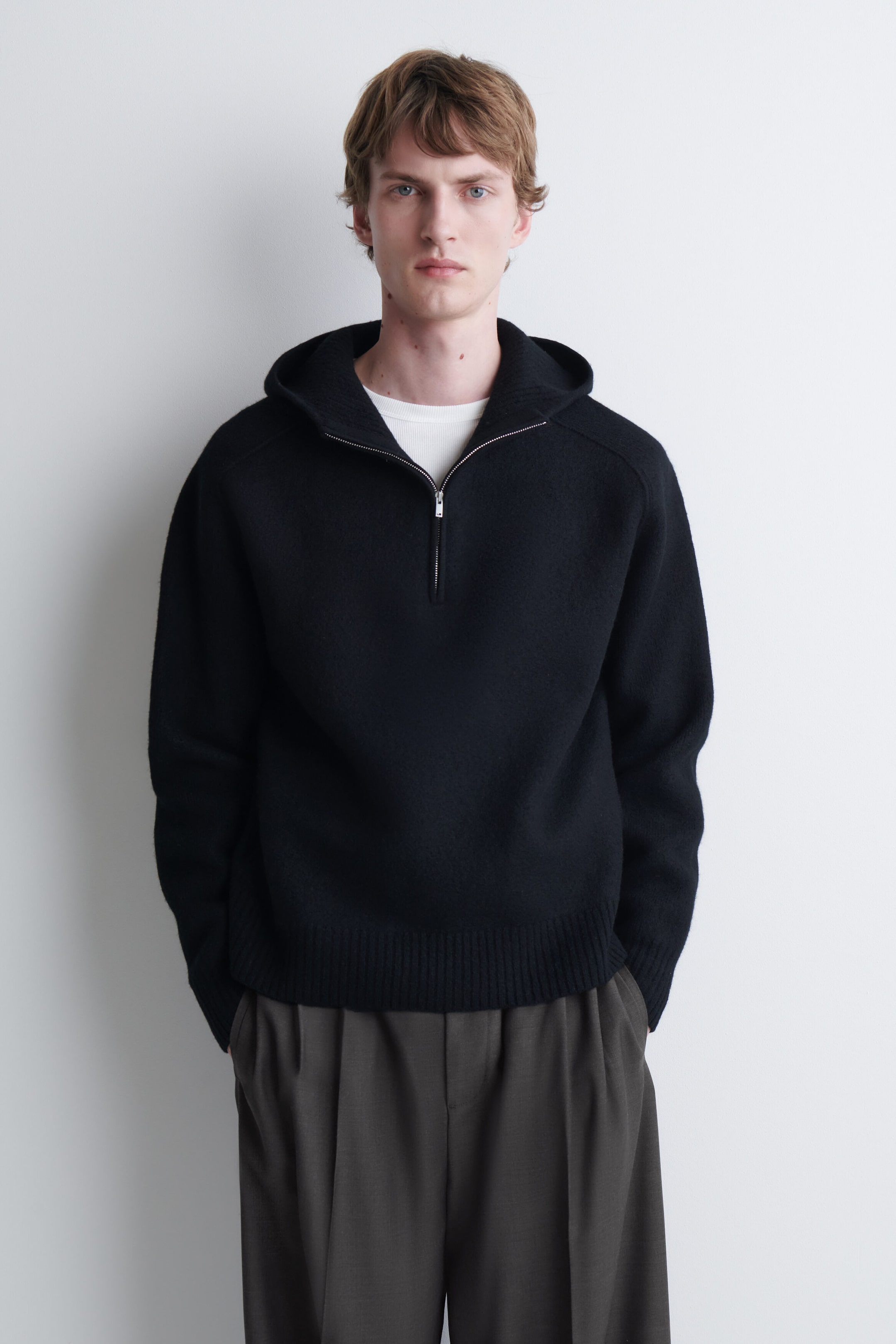 A black merino wool half-zip hoodie featuring a silver-tone zip, ribbed hem, and attached hood is being worn by the model. The knit hoodie is layered over a white T-shirt and paired with dark, pleated trousers. A black merino wool half-zip hoodie featuring a silver-tone zip, ribbed hem, and attached hood is being worn by the model. The knit hoodie is layered over a white T-shirt and paired with dark, pleated trousers.