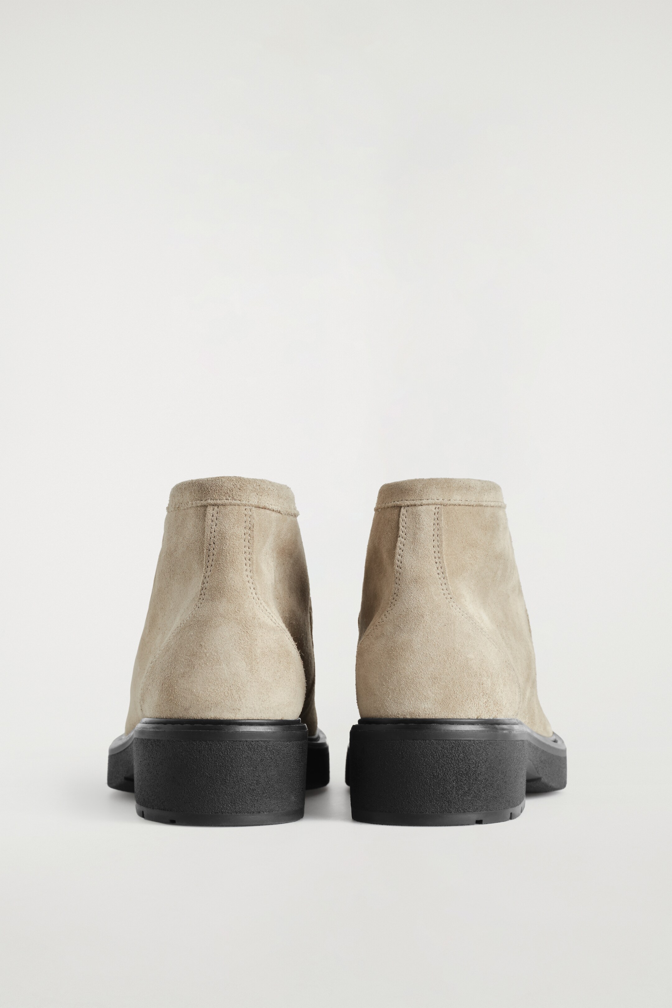 Grotere afbeelding bekijken: Een paar licht beige suède enkellaarsjes staan naar achteren gericht, met tonale stiksels langs de achterkant van de schachten en dikke zwarte contrasterende zolen.