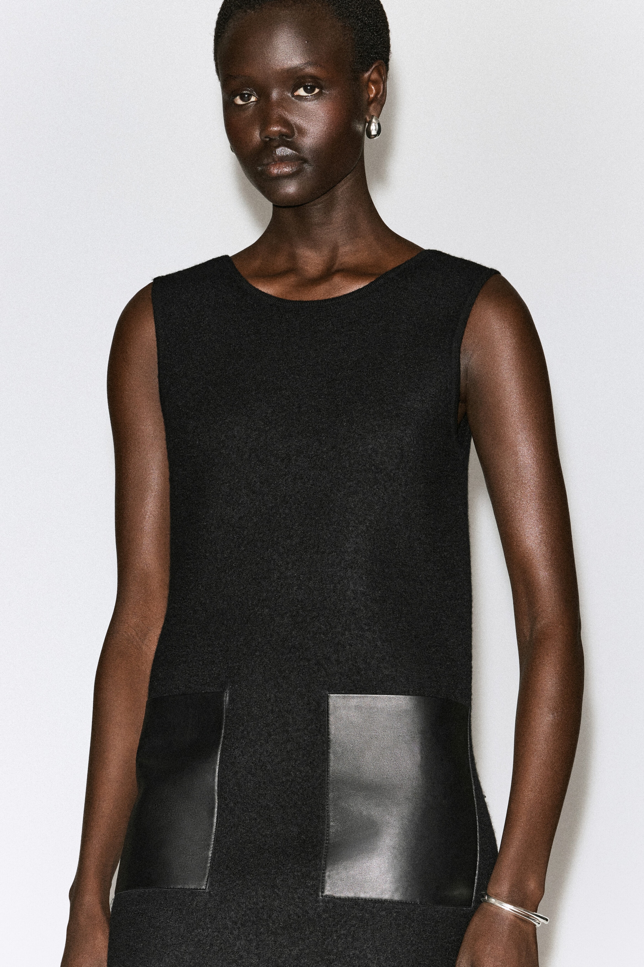 A model is wearing a sleeveless black mini dress crafted from textured boiled wool, featuring a round neckline and two large, smooth black leather patch pockets on the front. A model is wearing a sleeveless black mini dress crafted from textured boiled wool, featuring a round neckline and two large, smooth black leather patch pockets on the front.