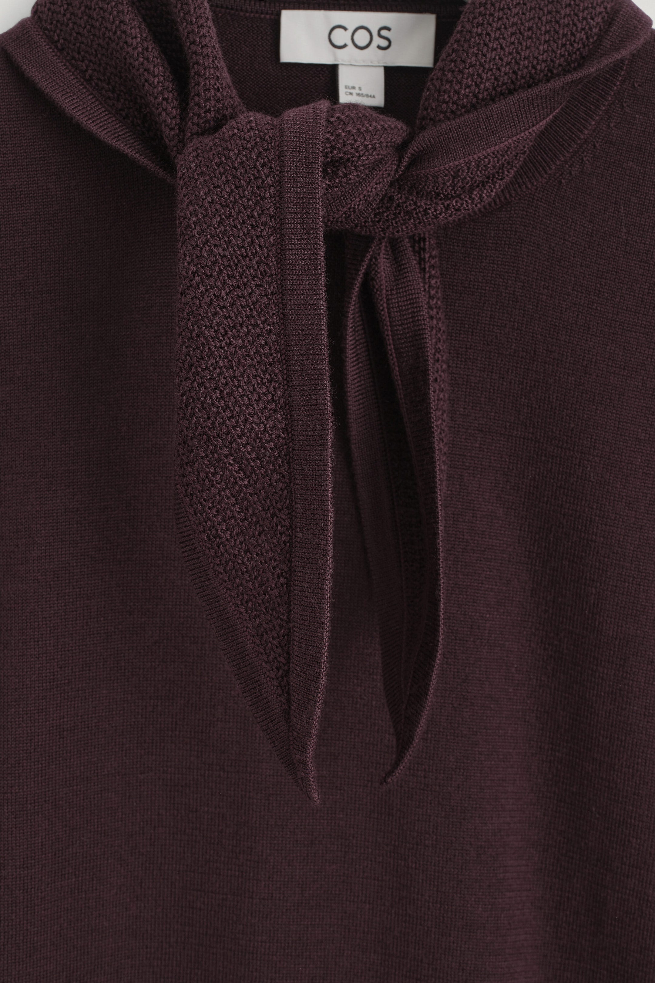 The scarf-detail merino wool-blend T-shirt features an integrated scarf tied loosely at the front neckline. The scarf displays a textured knit pattern with ribbed edges, contrasting with the smooth, fine-knit body of the deep plum-colored garment.