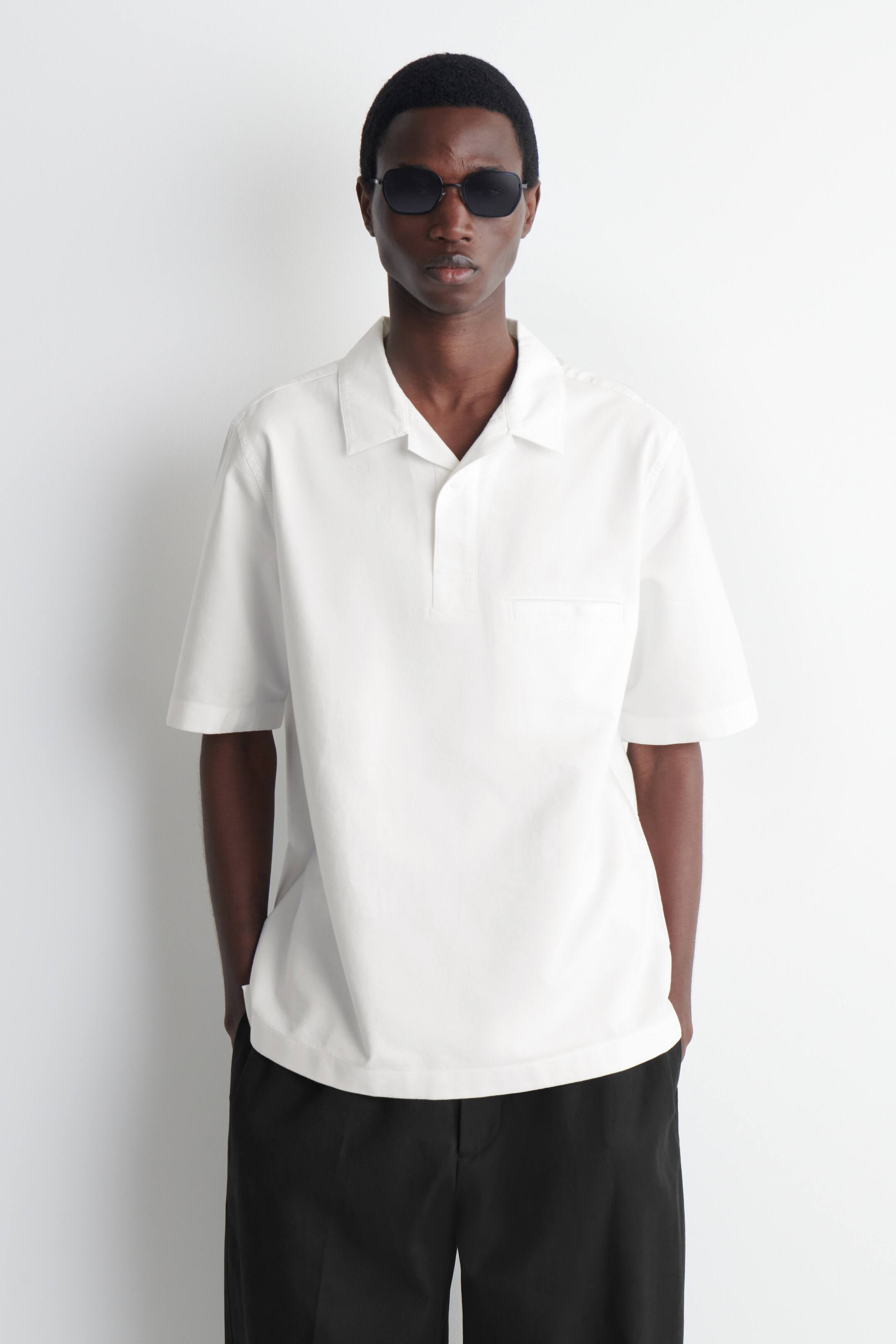 Ver imagem maior: Uma camisa branca de manga curta apresenta uma gola aberta, uma carcela curta e um único bolso de remendo no peito esquerdo. Exibe um corte descontraído com uma bainha reta.
