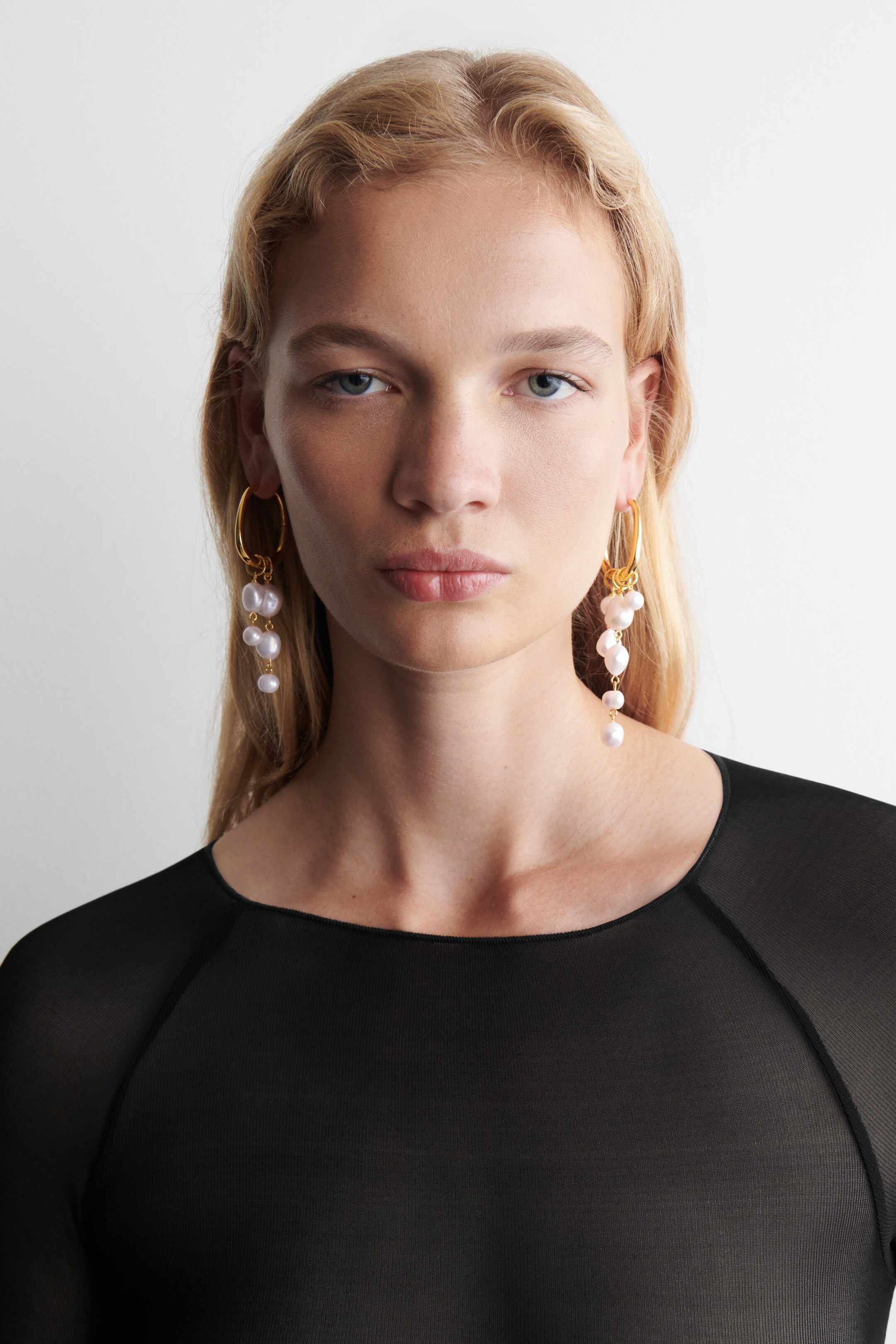 View larger image: A pair of gold-toned hoop earrings is featuring multiple white, irregularly shaped pearls that are dangling in cascading clusters, being presented from a frontal perspective.