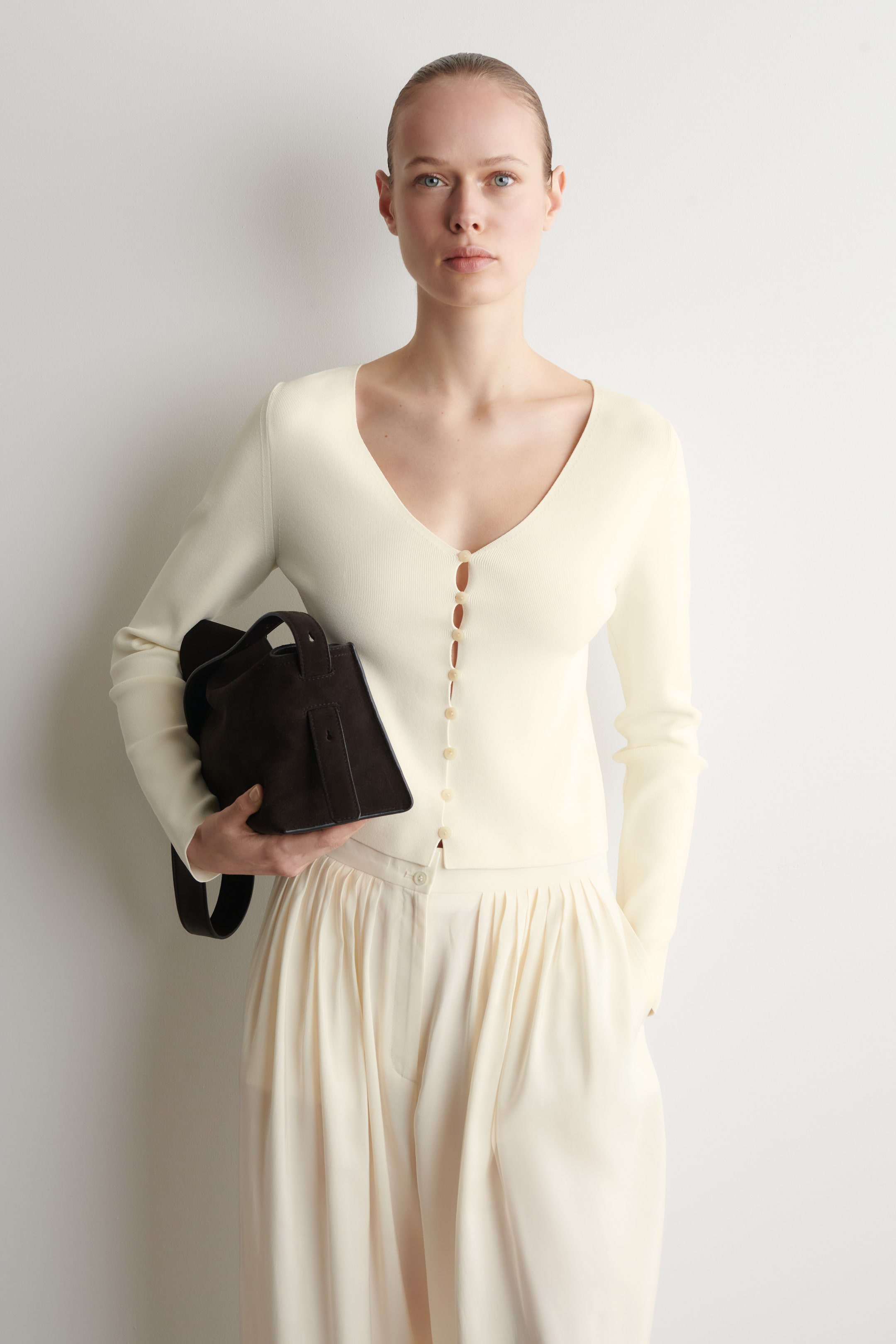 Visualizza immagine più grande: Un cardigan a maglia color bianco sporco presenta uno scollo a V e una chiusura frontale con piccoli bottoni chiari. È abbinato a pantaloni plissettati dello stesso colore bianco sporco, che mostrano un design a vita alta e gambe ampie.
