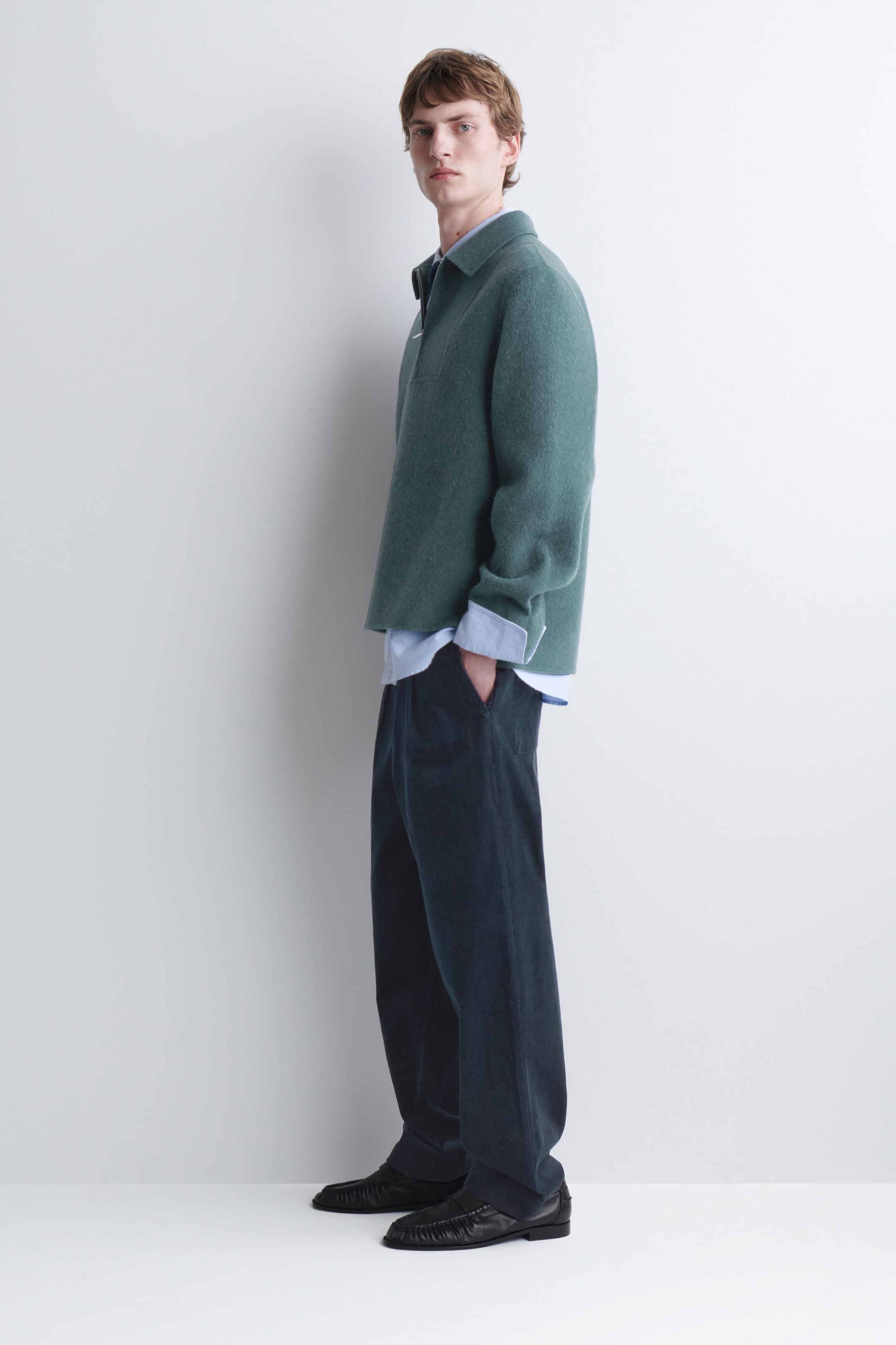 The model is wearing a green half-zip pullover with a pointed collar, layered over a light blue collared shirt with extended cuffs. The pullover is styled with dark wide-leg trousers and black leather loafers. The model is wearing a green half-zip pullover with a pointed collar, layered over a light blue collared shirt with extended cuffs. The pullover is styled with dark wide-leg trousers and black leather loafers.