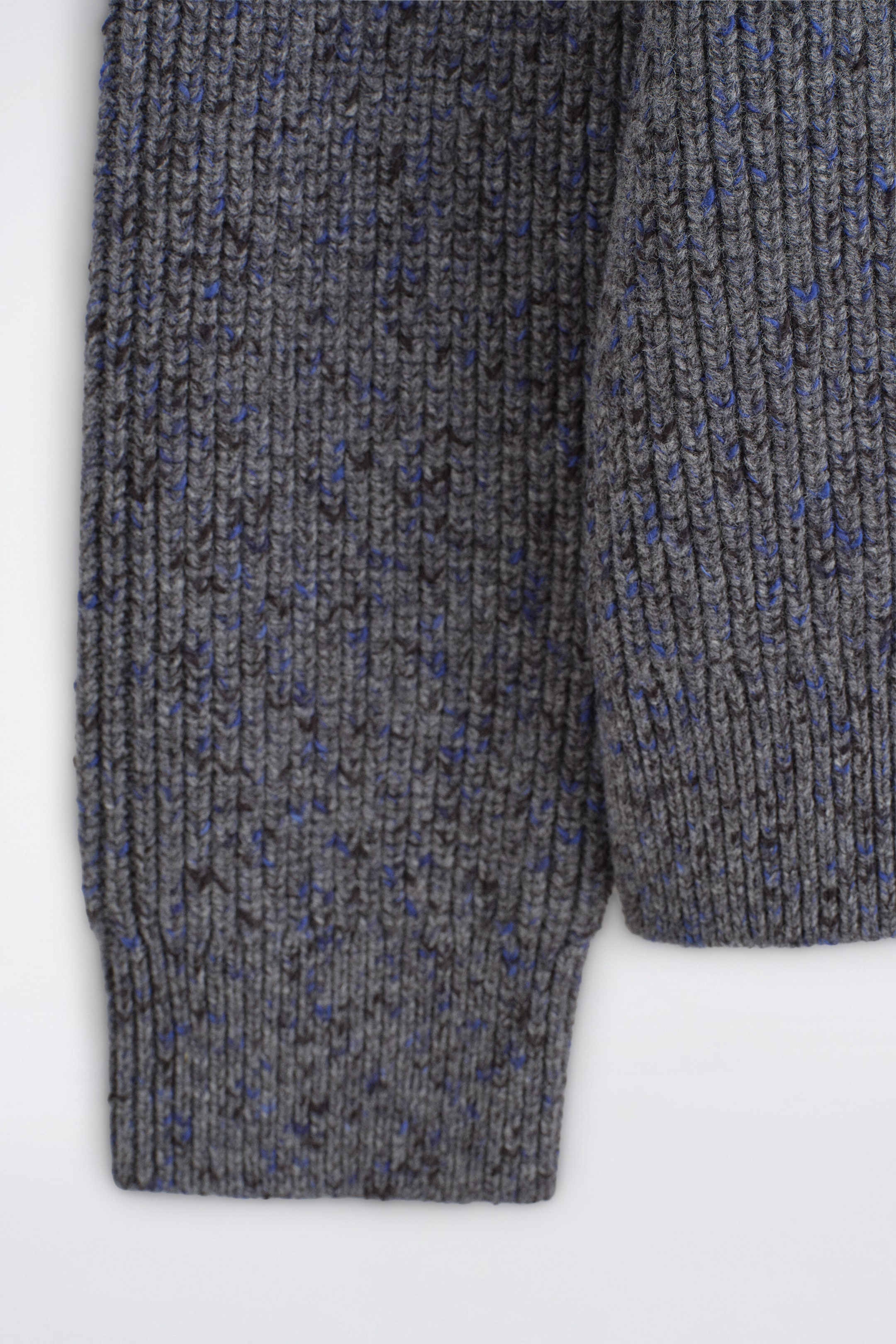 A close-up front view shows the thick-ribbed knit of a grey sweater featuring blue and dark brown flecks throughout the yarn. The design features a ribbed cuff on the sleeve and a matching ribbed hem. A close-up front view shows the thick-ribbed knit of a grey sweater featuring blue and dark brown flecks throughout the yarn. The design features a ribbed cuff on the sleeve and a matching ribbed hem.