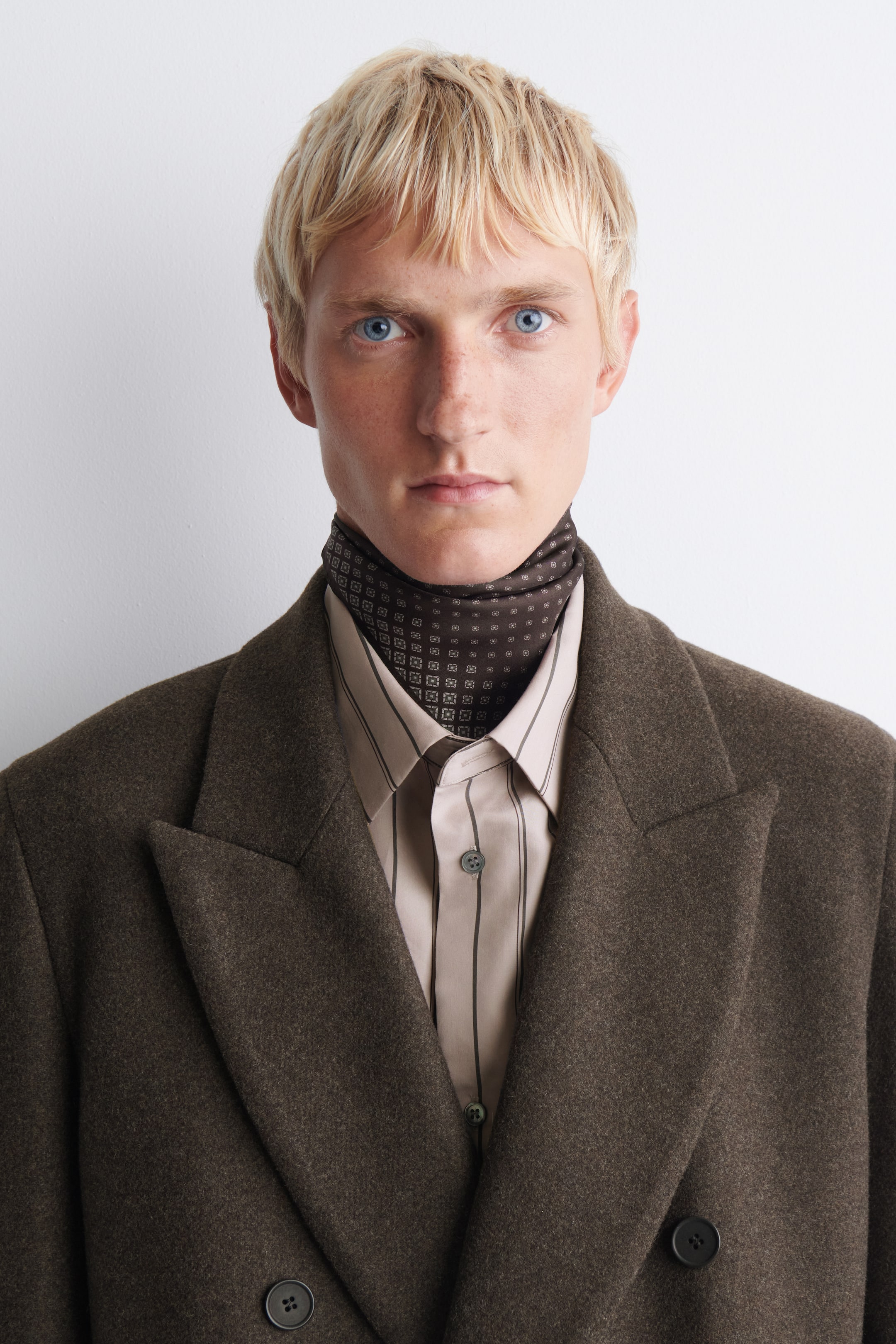 View larger image: A brown double-breasted wool coat features wide peak lapels and dark buttons in a front-facing view. The coat is layered over a beige striped button-down shirt and a dark brown patterned neck scarf.