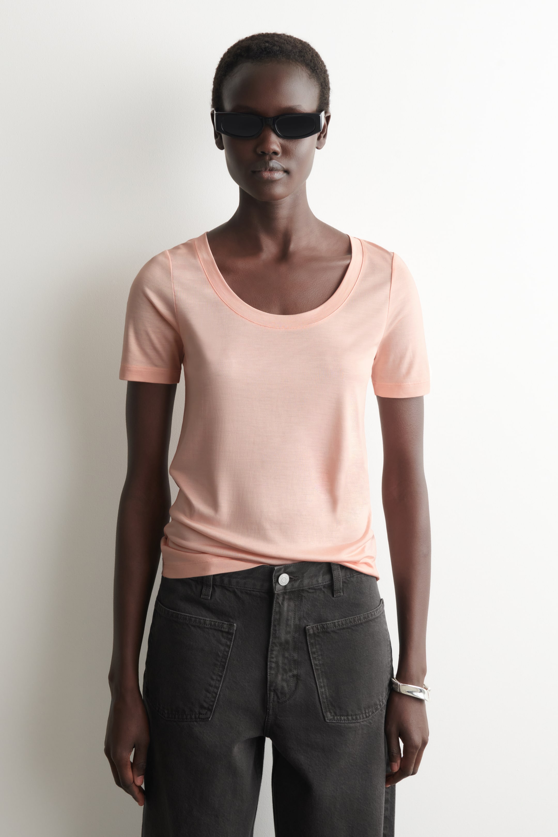 הצגת תמונה גדולה יותר: חולצת טי בצבע אפרסק בהיר מוצגת מהחזית, עם קו צוואר מעוגל בעדינות ושרוולים קצרים. היא מציגה גזרה צמודה ומוכנסת חלקית למכנסיים כהים.