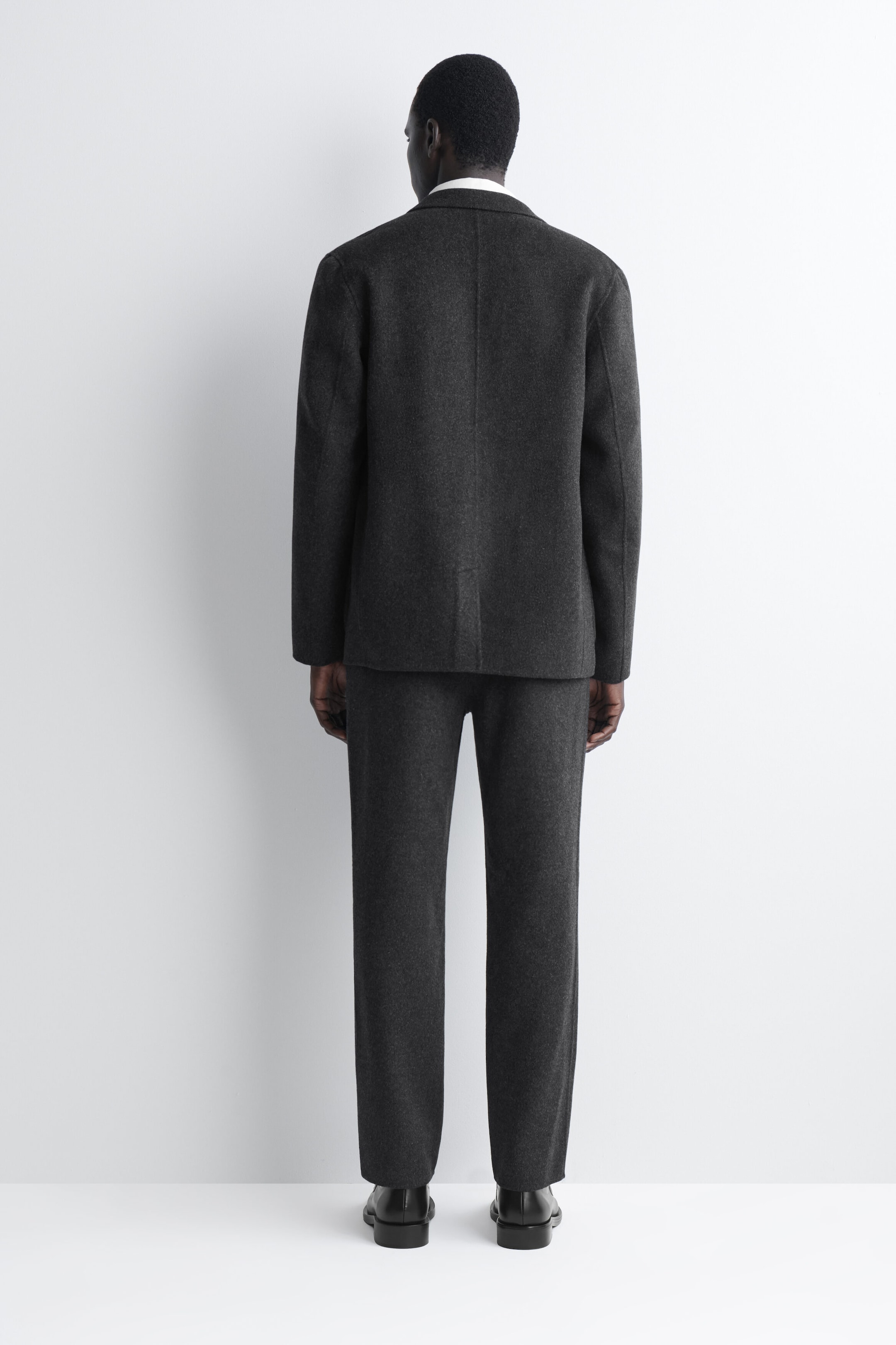 These dark grey, straight-leg trousers are shown from the back, featuring a relaxed fit in a textured wool-blend fabric. They are styled with a matching unstructured blazer and black dress shoes, highlighting their clean, unstructured drape. These dark grey, straight-leg trousers are shown from the back, featuring a relaxed fit in a textured wool-blend fabric. They are styled with a matching unstructured blazer and black dress shoes, highlighting their clean, unstructured drape.