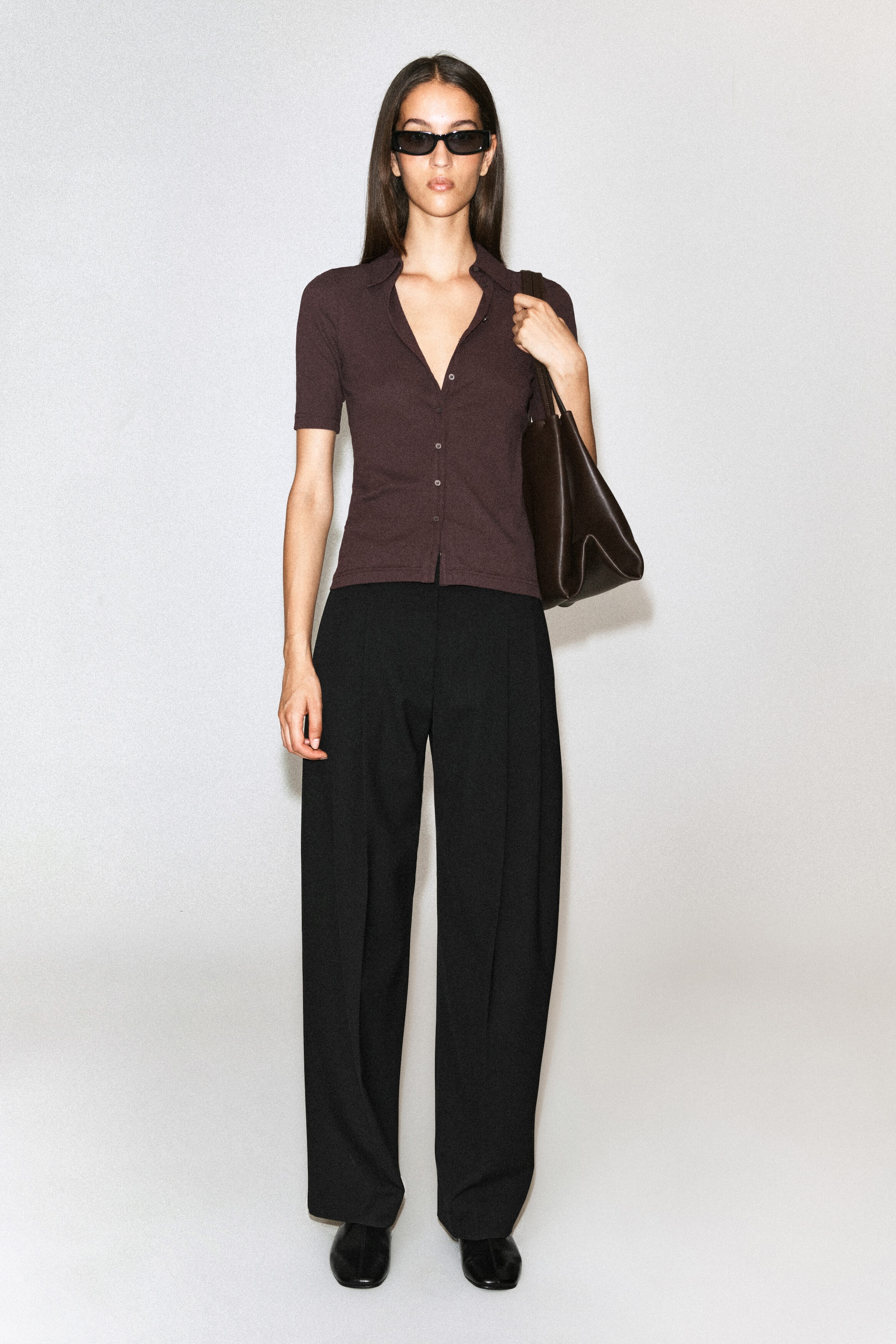 A dark brown, short-sleeved cotton jersey shirt with a collar and button-front detail is worn by the model. The slim-fit shirt is styled with black wide-leg trousers, and a dark brown leather shoulder bag is carried. A dark brown, short-sleeved cotton jersey shirt with a collar and button-front detail is worn by the model. The slim-fit shirt is styled with black wide-leg trousers, and a dark brown leather shoulder bag is carried.