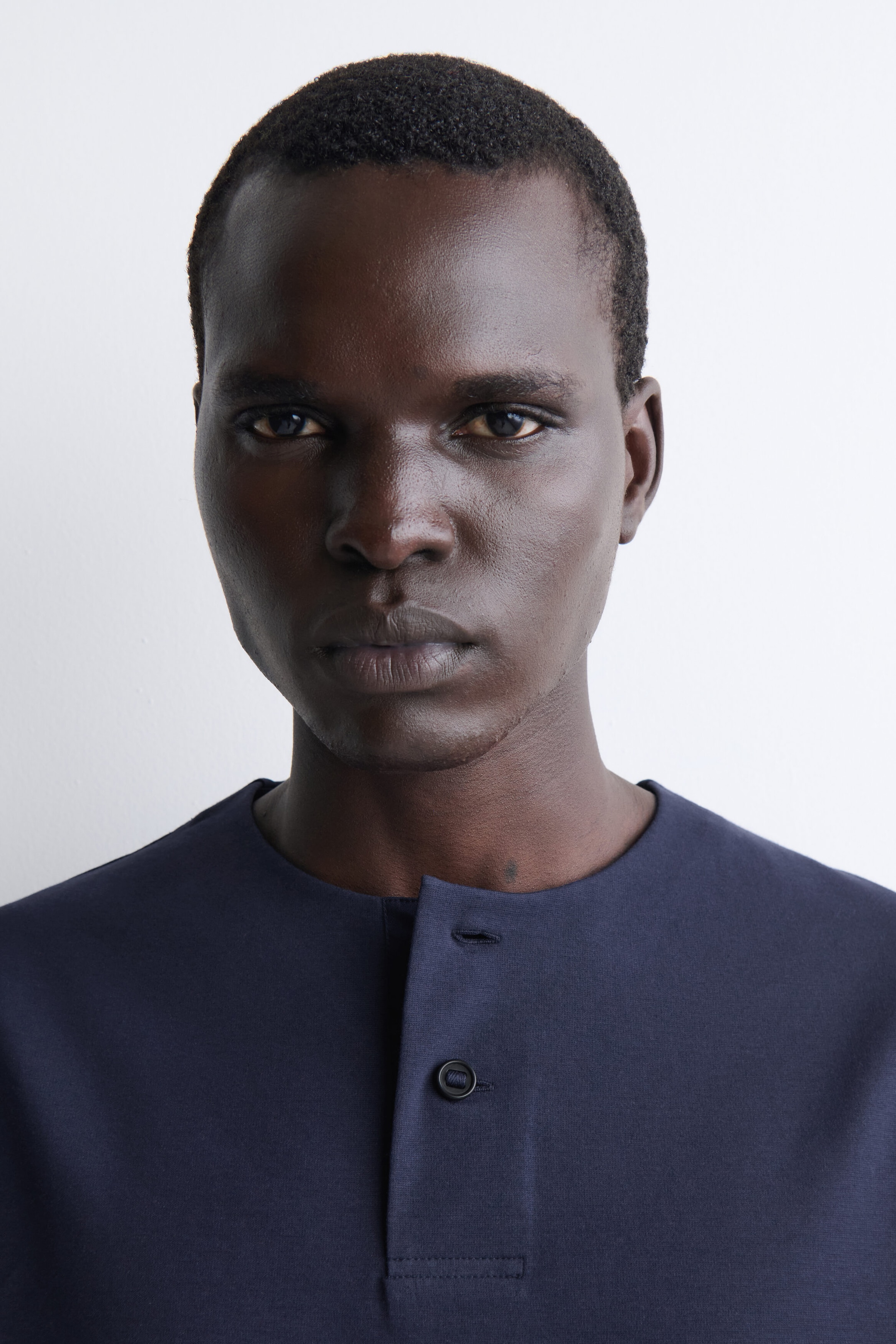 Un t-shirt henley oversize en jersey de coton bleu marine est présenté en gros plan, vue de face de la partie supérieure, mettant en valeur son col rond épuré. Le modèle comprend une patte de boutonnage partielle avec un seul bouton foncé fermé et un détail de boutonnière ouverte au col.