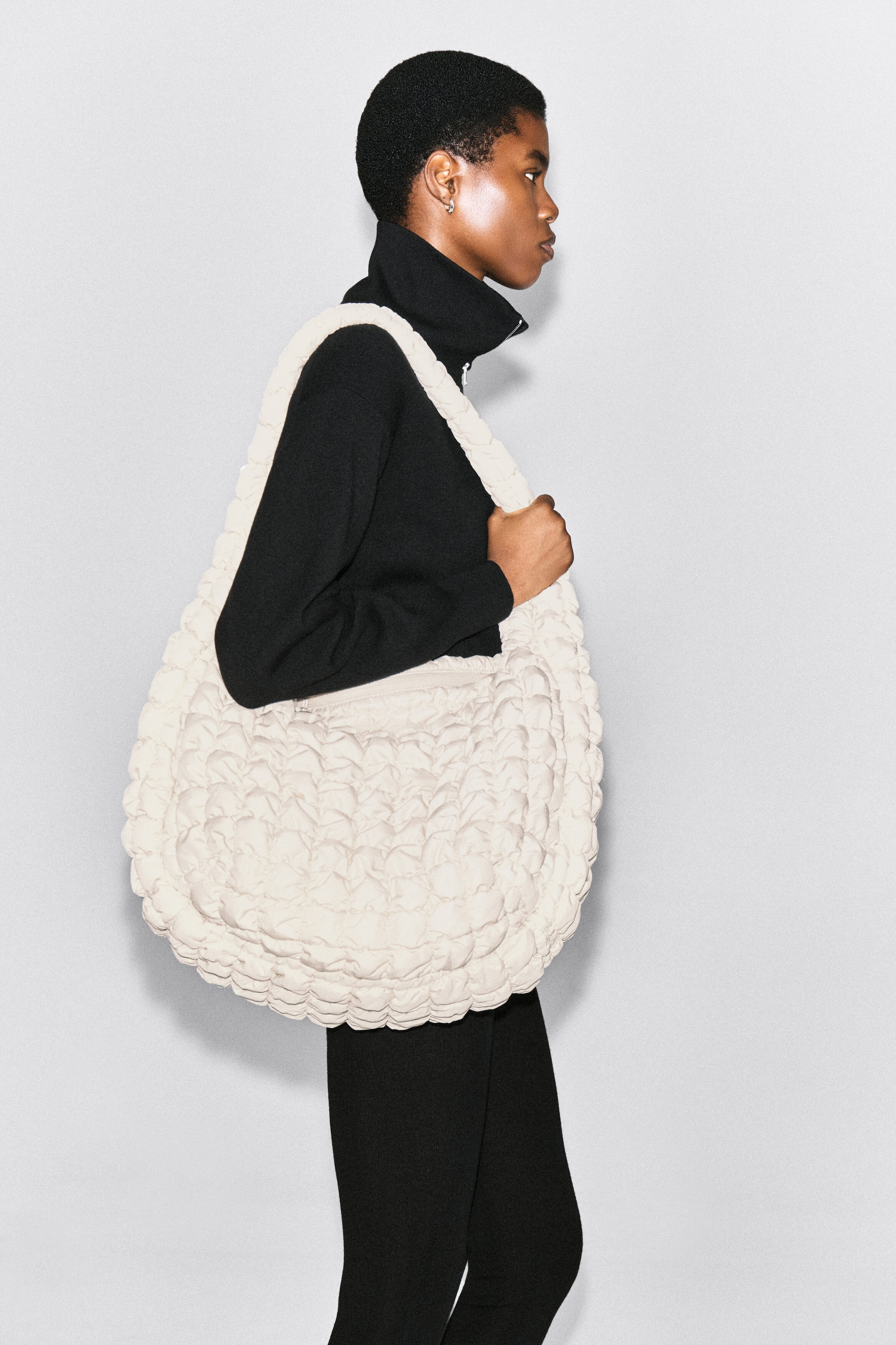 An oversized quilted crossbody bag in off-white is worn over the model’s shoulder as she holds the wide strap. The bag features a distinctive puckered, quilted texture that contrasts with the model’s black high-neck top and trousers. An oversized quilted crossbody bag in off-white is worn over the model’s shoulder as she holds the wide strap. The bag features a distinctive puckered, quilted texture that contrasts with the model’s black high-neck top and trousers.