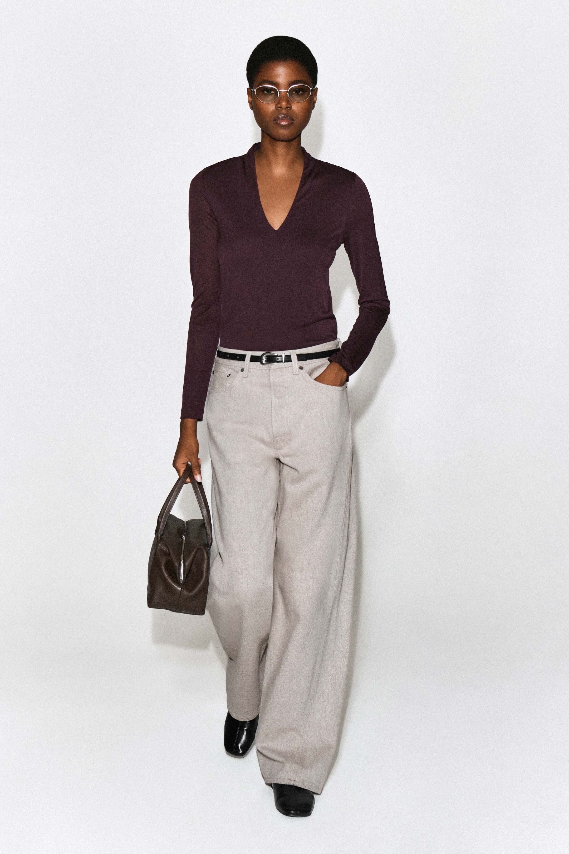 A dark plum, long-sleeved top with a deep V-neckline and a draped, shawl-like collar is worn by the model. The slim-fit top is tucked into light-colored, wide-leg trousers styled with a thin black belt. A dark plum, long-sleeved top with a deep V-neckline and a draped, shawl-like collar is worn by the model. The slim-fit top is tucked into light-colored, wide-leg trousers styled with a thin black belt.