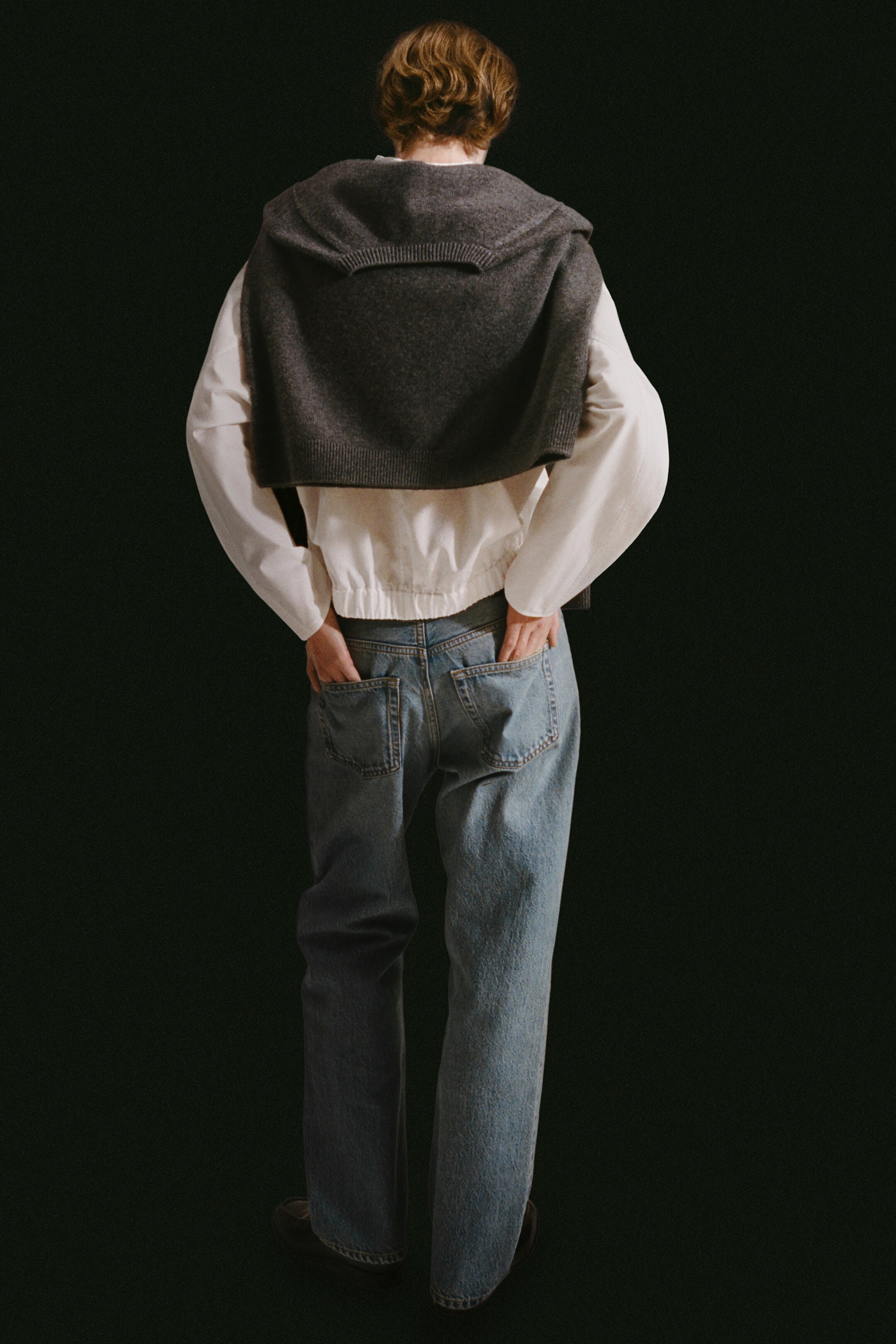 Größeres Bild anzeigen: Eine weiße Bluse aus Baumwollpopeline wird getragen, die eine sanft abgerundete Silhouette mit einem elastischen Saum am Rücken aufweist. Sie wird mit hellblauen Jeans mit geradem Bein kombiniert, die sichtbare aufgesetzte Gesäßtaschen haben. Ein dunkelgrauer Pullover aus Merinowolle ist über die Schultern drapiert, wobei der gerippte V-Ausschnitt ein breites, flaches Kragendetail am Rücken bildet.