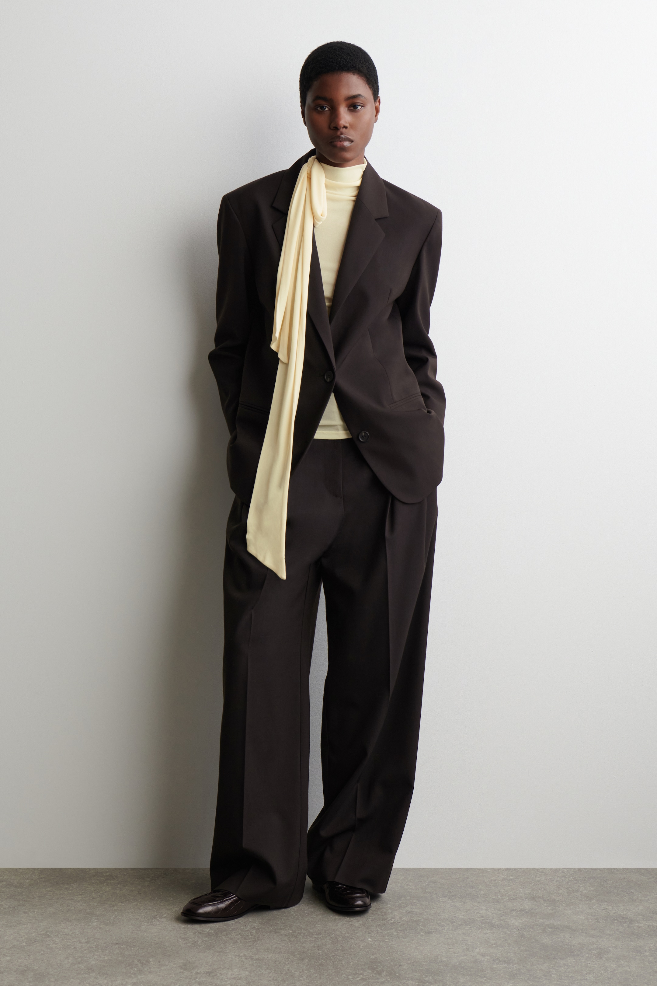 Agrandir l'image: Un costume marron foncé est présenté, composé d'un blazer à coupe décontractée avec des épaules structurées proéminentes, des revers crantés et des poches à rabat, assorti à un pantalon large avec des plis à l'avant. Un haut à col montant jaune clair est superposé sous le blazer, avec une longue écharpe coordonnée drapée sur le costume.