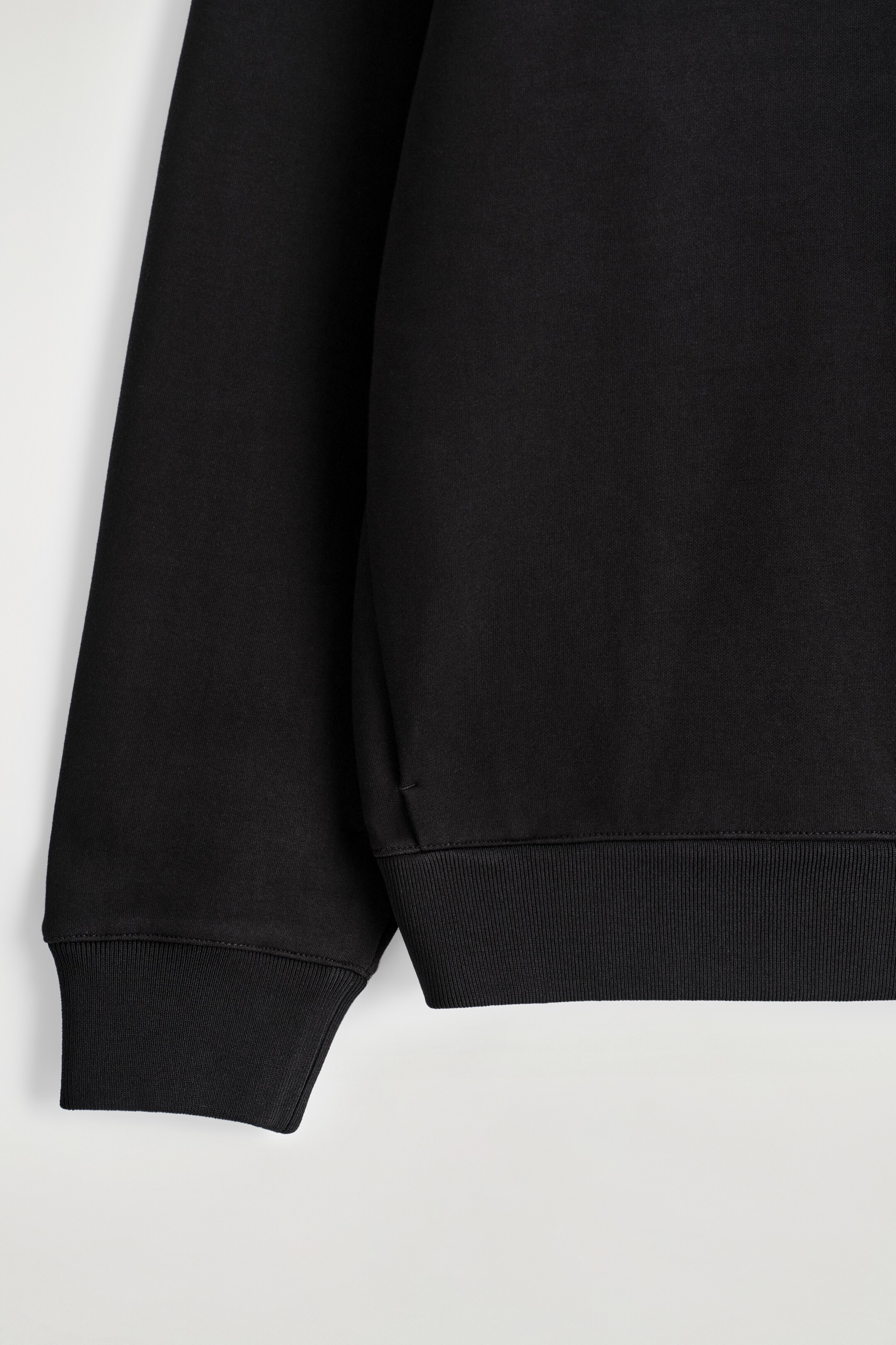 Agrandir l'image: Un sweat-shirt noir est montré en gros plan, avec un tissu lisse et dense. Sa manche longue se termine par un poignet côtelé, et le bas du corps se termine par un large ourlet côtelé, avec un petit détail de boucle visible près de la couture latérale.