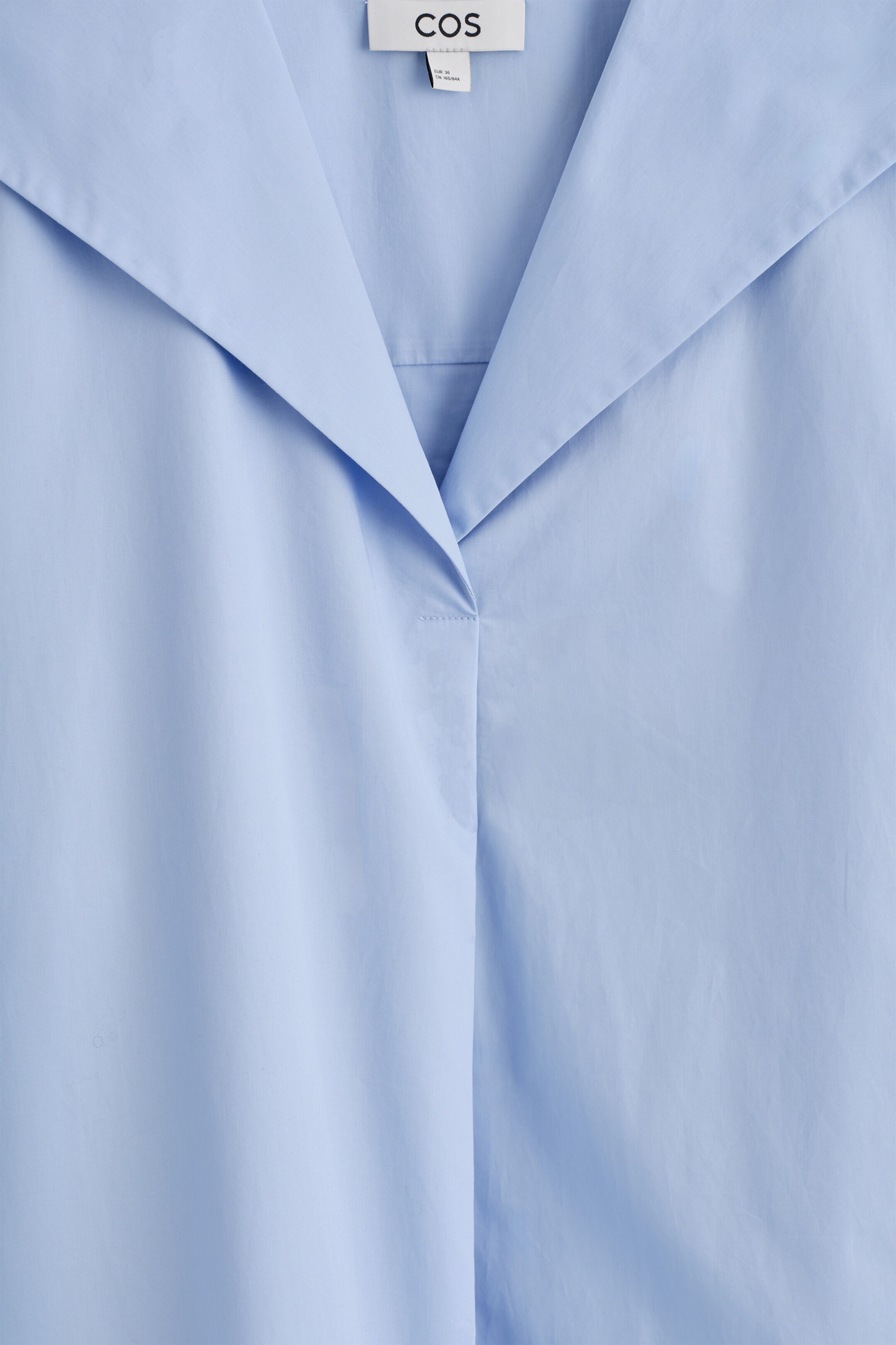 Agrandir l'image: Une robe bleu clair présente un large col marin plat qui s'étend sur les épaules. Le tissu en popeline de coton frais forme un décolleté en V à l'avant, avec une étiquette de marque 'COS' visible en haut.