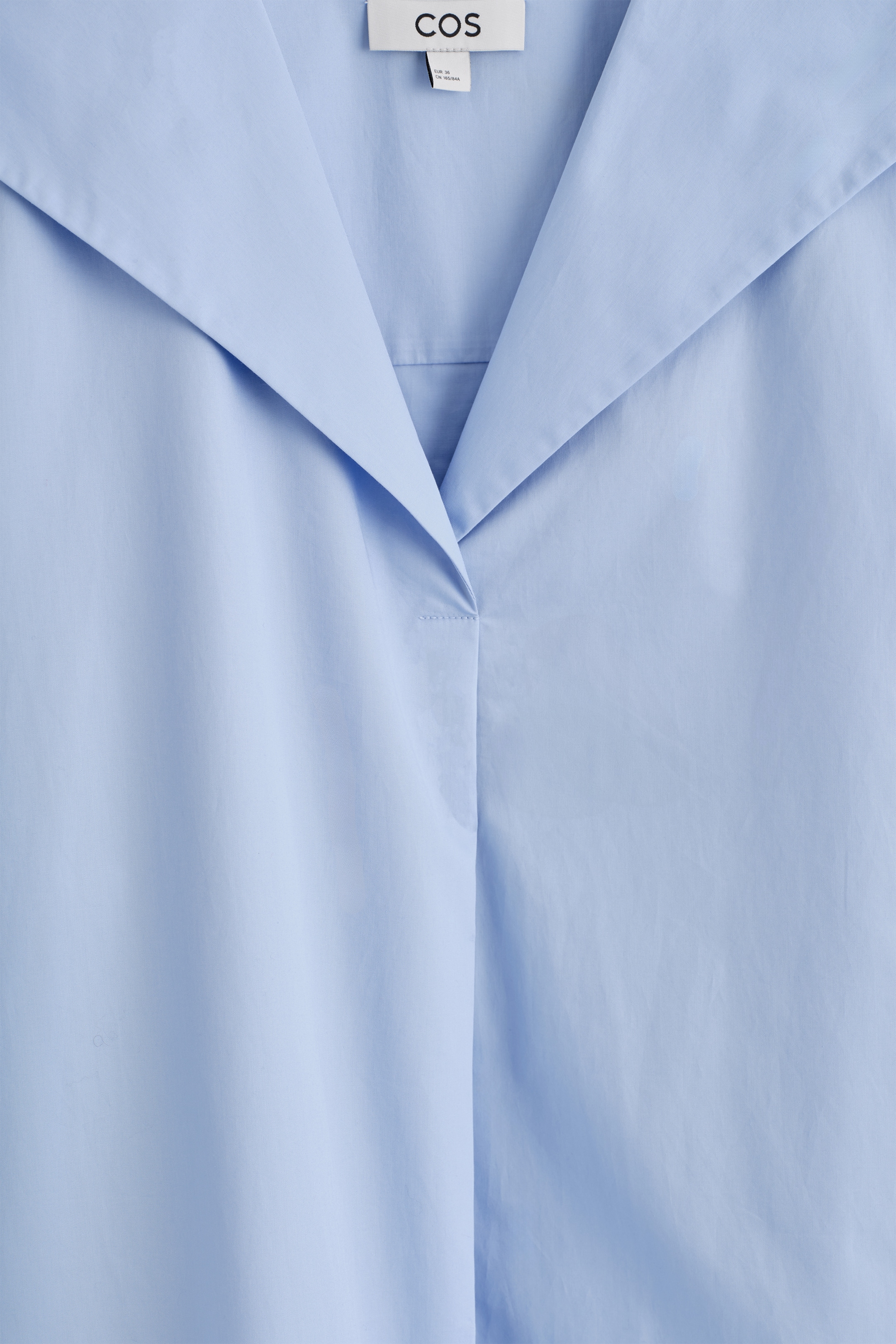 Ingrandisci l'immagine: Un abito azzurro chiaro presenta un ampio colletto da marinaio piatto che si estende sulle spalle. Il tessuto di cotone popeline croccante forma uno scollo a V sul davanti, con un'etichetta del marchio 'COS' visibile in alto.