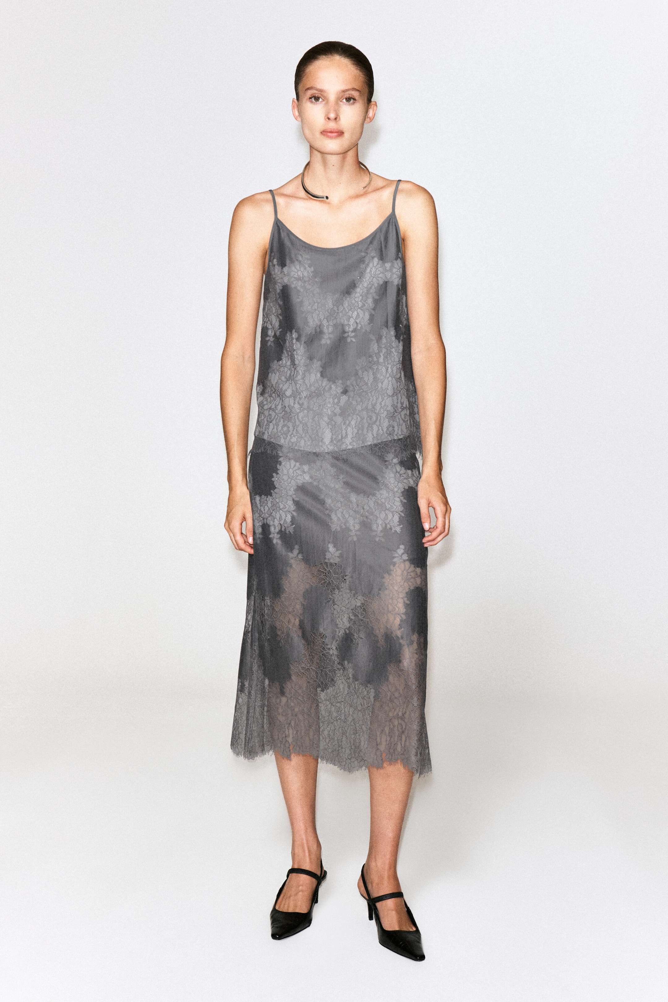 A grey lace-overlay camisole top featuring thin spaghetti straps and a floral lace pattern is being worn by the model. The top is styled with a matching grey midi skirt, a silver collar necklace, and black heels. A grey lace-overlay camisole top featuring thin spaghetti straps and a floral lace pattern is being worn by the model. The top is styled with a matching grey midi skirt, a silver collar necklace, and black heels.
