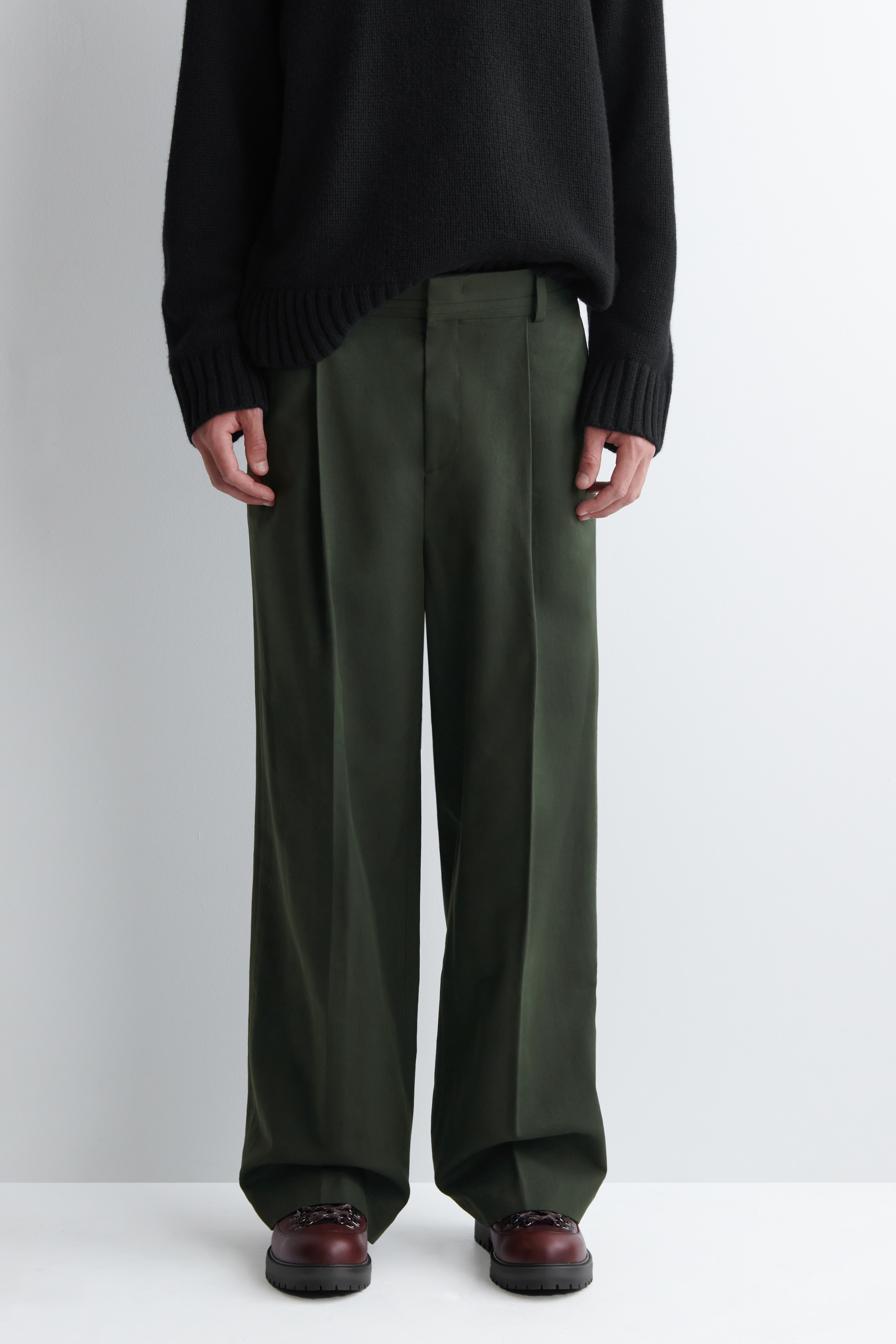 Ingrandisci l'immagine: Pantaloni a gamba larga verde scuro sono presentati dal FRONTE, con doppie pieghe in vita, passanti per cintura e una piega distintiva che corre lungo il centro di ciascuna gamba.