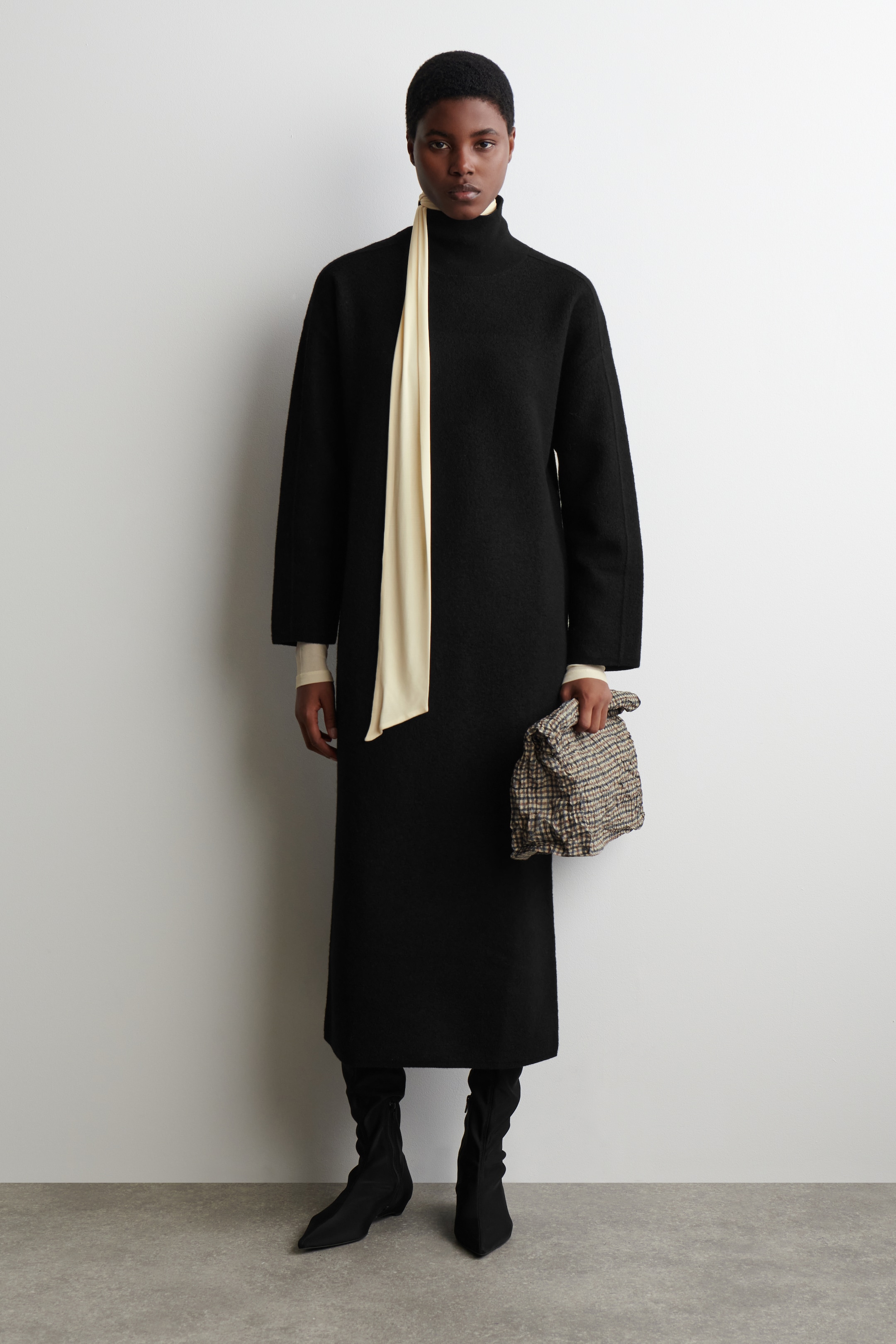 View larger image: A black, maxi-length dress is presenting a high neck and wide, long sleeves, displaying a straight, relaxed fit from a front-facing angle. A cream long-sleeved top is layering beneath the dress, with a coordinating cream scarf draping vertically from the neckline, while a textured clutch bag is being held and black pointed-toe ankle boots are visible.