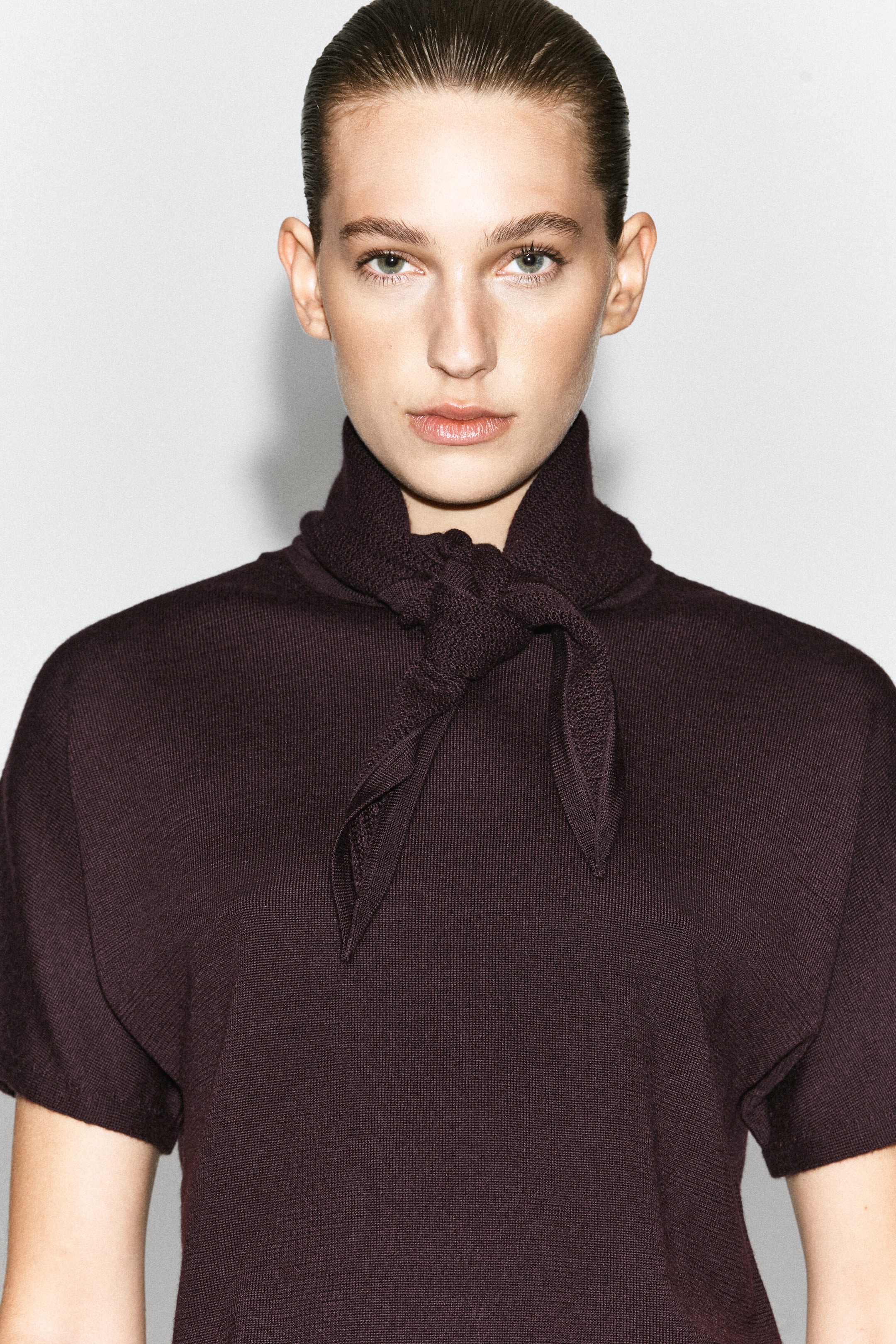 The model is wearing a dark burgundy merino wool-blend T-shirt. This short-sleeved garment features a high collar with an integrated matching knit scarf detail tied in a knot at the front.