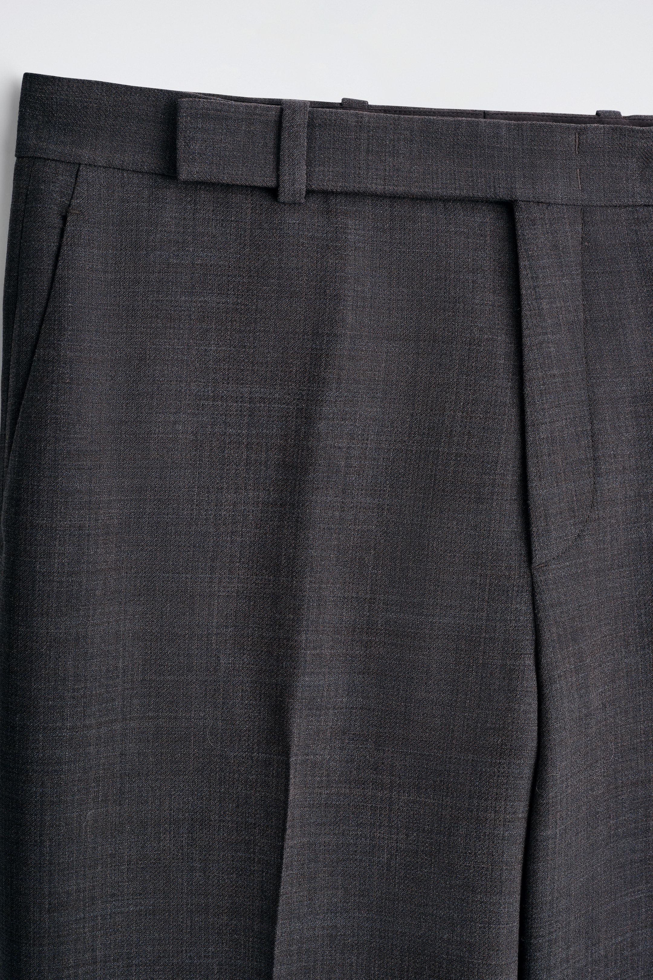 Agrandir l'image: Une paire de pantalons gris foncé est présentée de face, avec un tissage texturé subtil et une ceinture visible avec des passants pour ceinture. Une ouverture de poche latérale est visible à gauche, et un seul pli descend verticalement le long de la jambe droite.