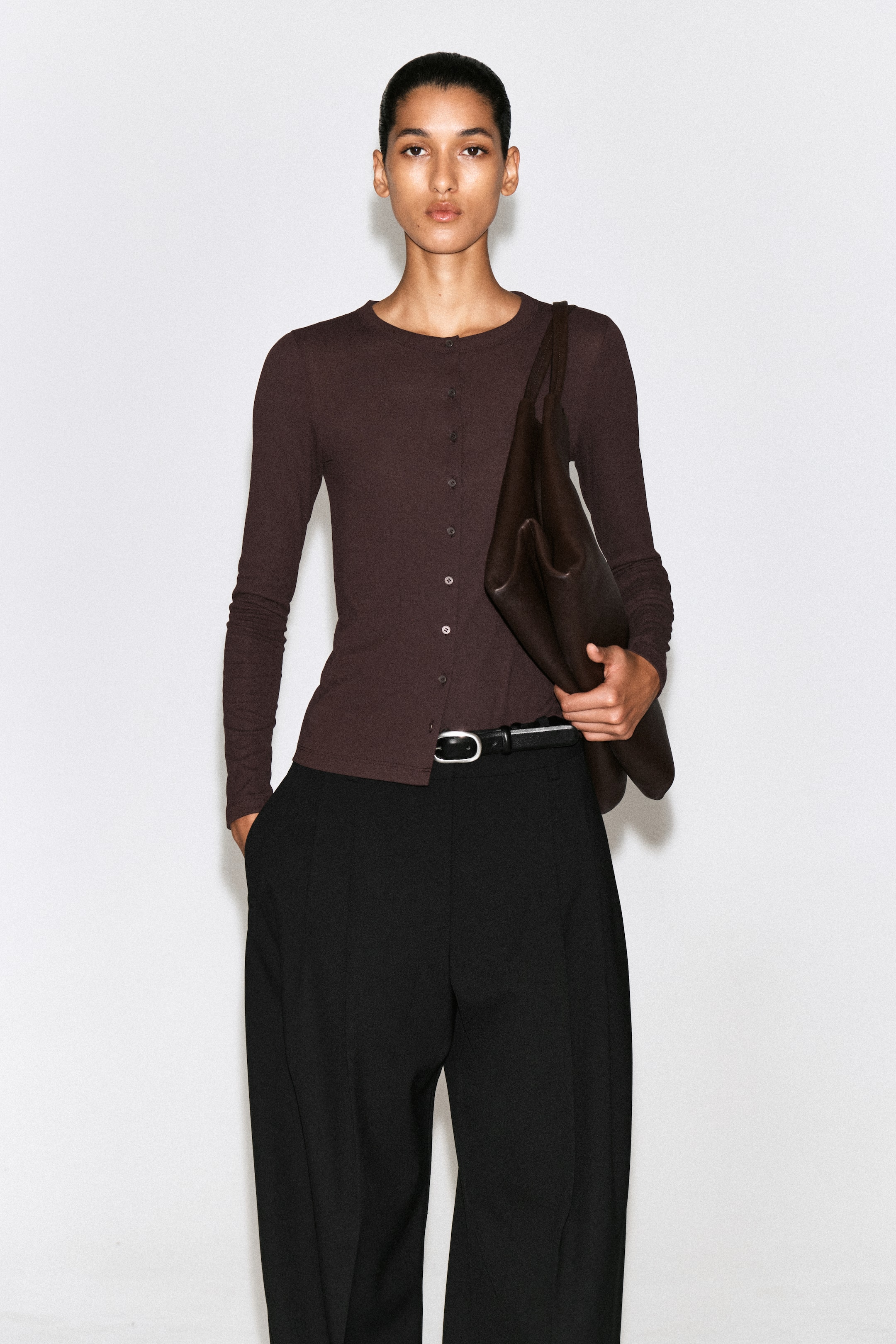 Visualizza l'immagine ingrandita: Un cardigan top in jersey marrone scuro mostra una silhouette slim fit con girocollo e maniche lunghe, ripreso frontalmente. Il top presenta una chiusura frontale con bottoni ed è abbinato con una parziale rimboccatura in pantaloni neri con cintura.