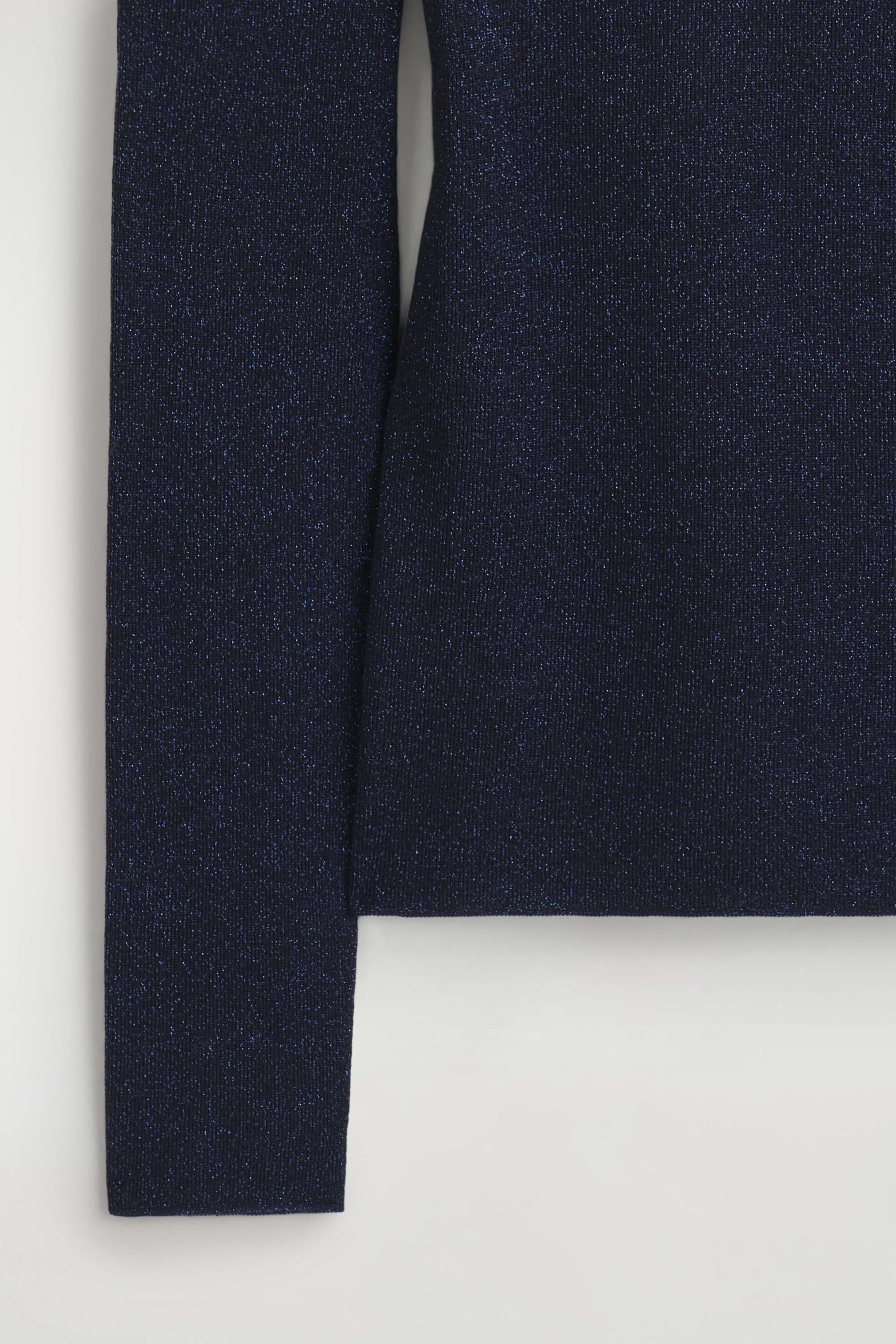 The sparkly seamless wool funnel-neck top features a dark navy blue, fine-gauge knit fabric woven with shimmering blue metallic threads. This close-up view shows the straight hemline of the body alongside a long, fitted sleeve with clean, seamless edges.