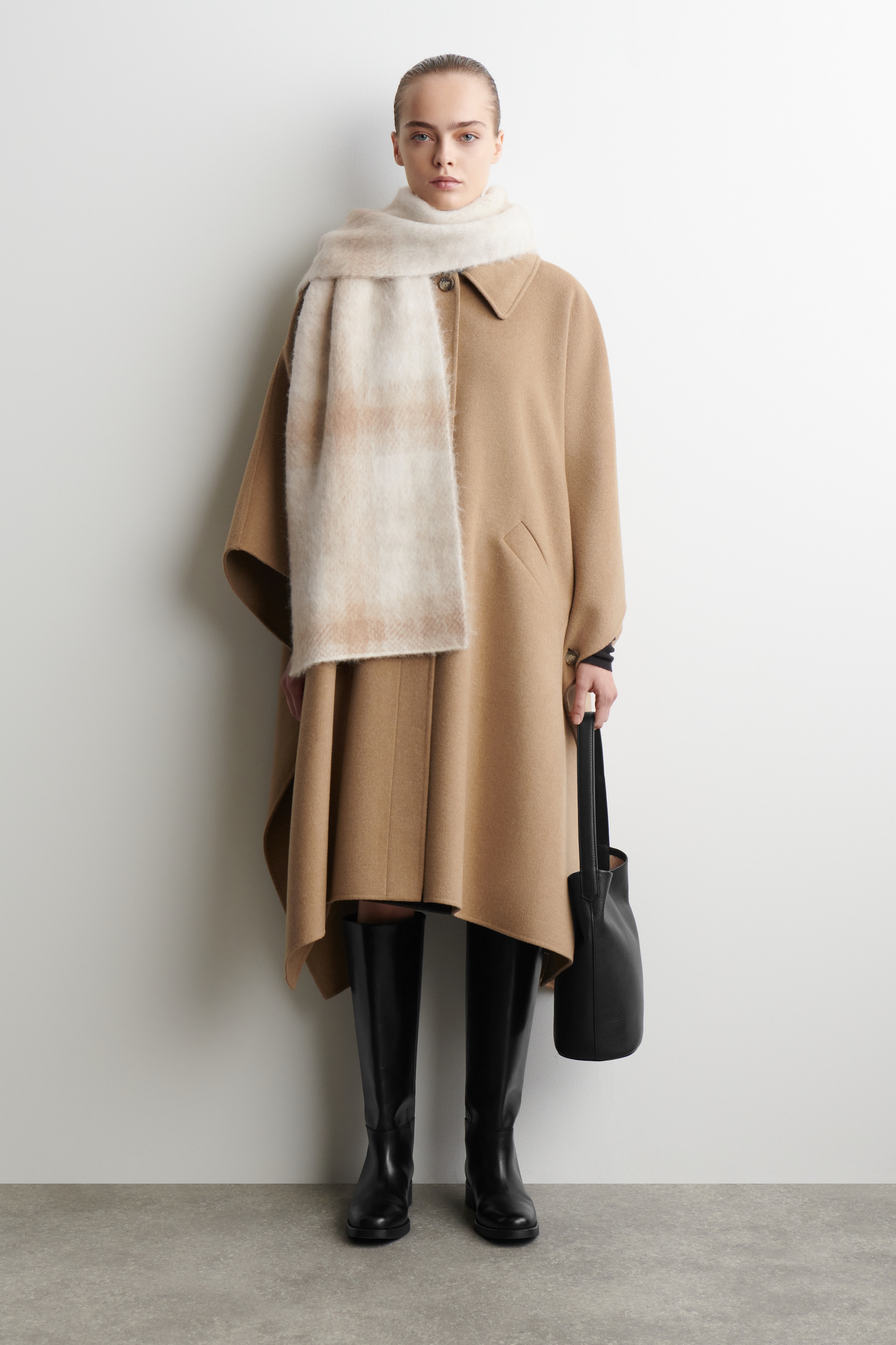 Agrandir l'image: Un manteau cape couleur camel est présenté de face, avec un col pointu, une fermeture à bouton unique au niveau du cou et des poches en biais, affichant une silhouette fluide. Une écharpe à carreaux crème et marron clair est drapée sur ses épaules, tandis qu'un sac cabas en cuir noir est tenu contre son côté, et des bottes noires jusqu'aux genoux se montrent sous son ourlet.