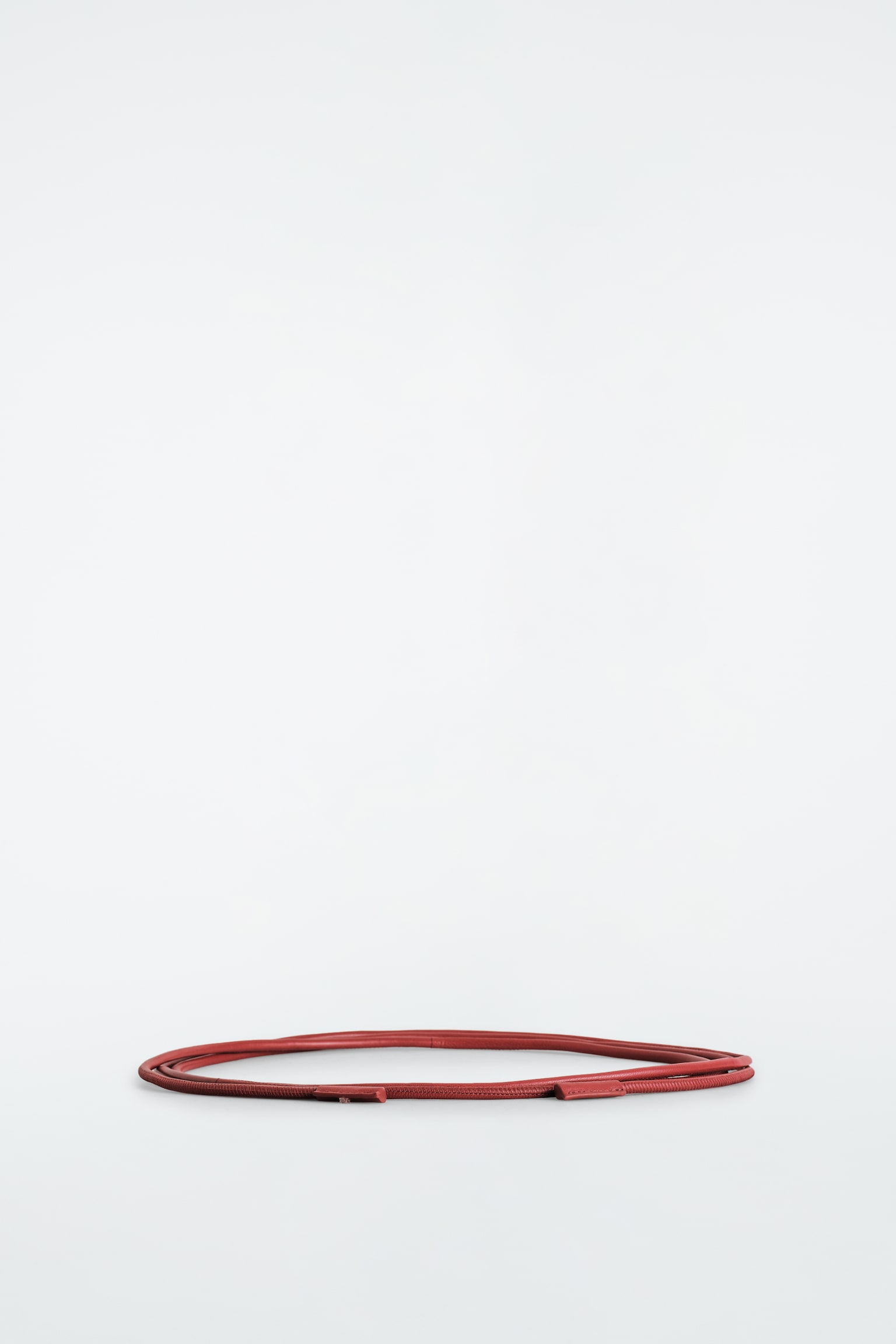 La ceinture en corde de cuir est posée enroulée dans une boucle horizontale plate, mettant en valeur son design fin et arrondi dans une couleur brun rougeâtre. De fins détails de couture longent toute sa longueur, avec de petits embouts en cuir assortis terminant les extrémités. - 1