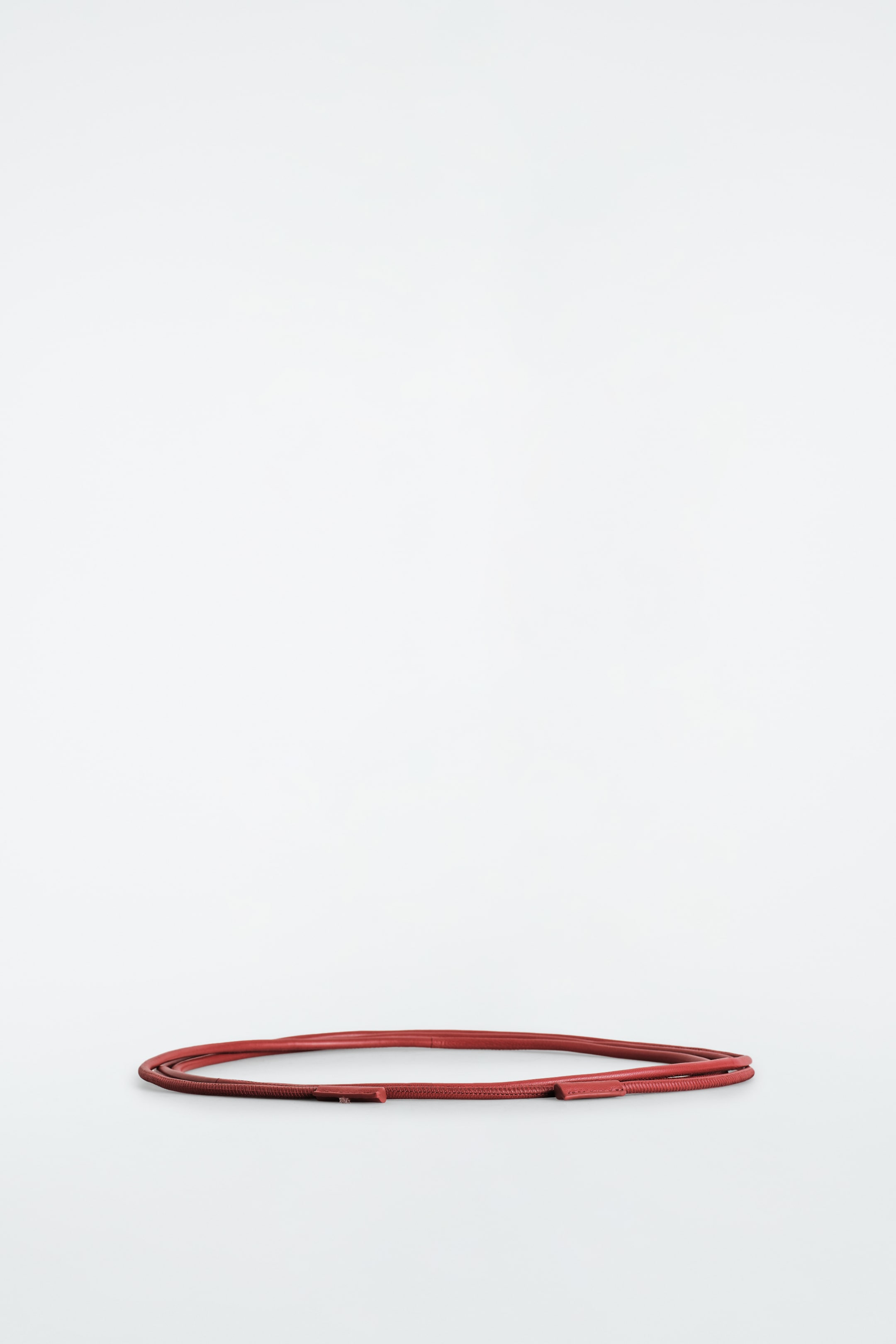 Agrandir l'image: La ceinture en corde de cuir est posée enroulée dans une forme ovale lâche, mettant en valeur sa fine construction en cuir rouge arrondi. La ceinture présente des embouts en cuir cousus aux deux extrémités.